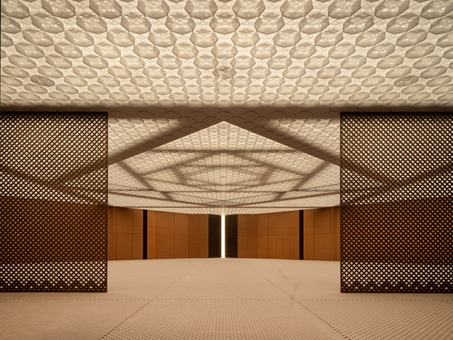 A prayer hall with wooden walls and a ceiling with integrated lighting. The ceiling features layers of patterned, perforated aluminum creating a soft ethereal effect. The structural system of steel beams in interlocking squares is visible above the ceiling.
