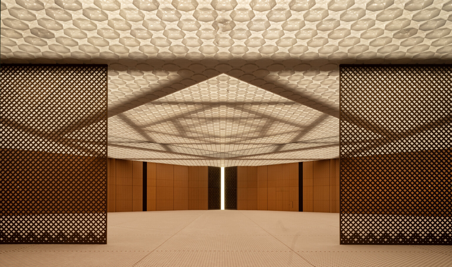 A prayer hall with wooden walls and a ceiling with integrated lighting. The ceiling features layers of patterned, perforated aluminum creating a soft ethereal effect. The structural system of steel beams in interlocking squares is visible above the ceiling.