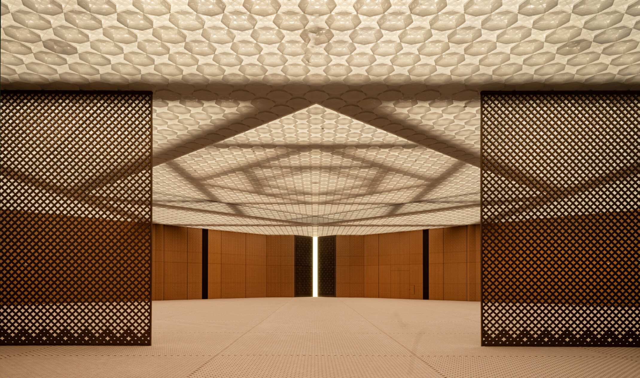 A prayer hall with wooden walls and a ceiling with integrated lighting. The ceiling features layers of patterned, perforated aluminum creating a soft ethereal effect. The structural system of steel beams in interlocking squares is visible above the ceiling.