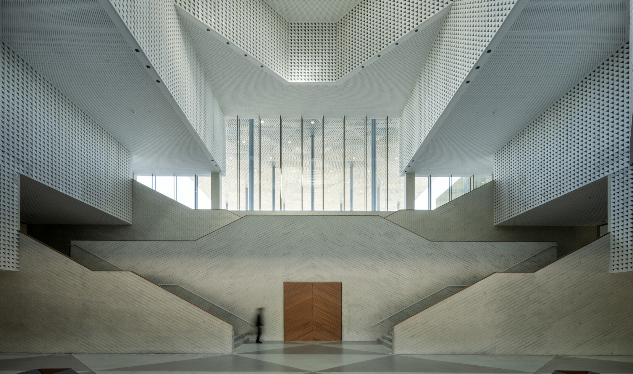 Atrium with perforated walls rising multiple stories around a dramatic angular staircase. Natural light floods through the oculus above and tall glass windows. Visitors move through the bright, open space, some seated in the foreground.