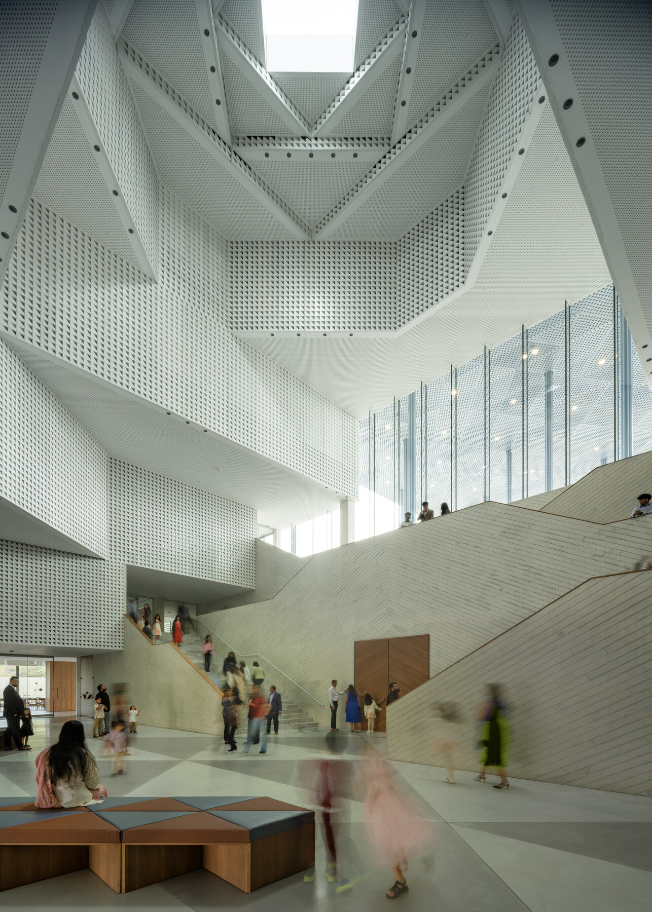 Atrium with perforated walls rising multiple stories around a dramatic angular staircase. Natural light floods through the oculus above and tall glass windows. Visitors move through the bright, open space, some seated in the foreground.