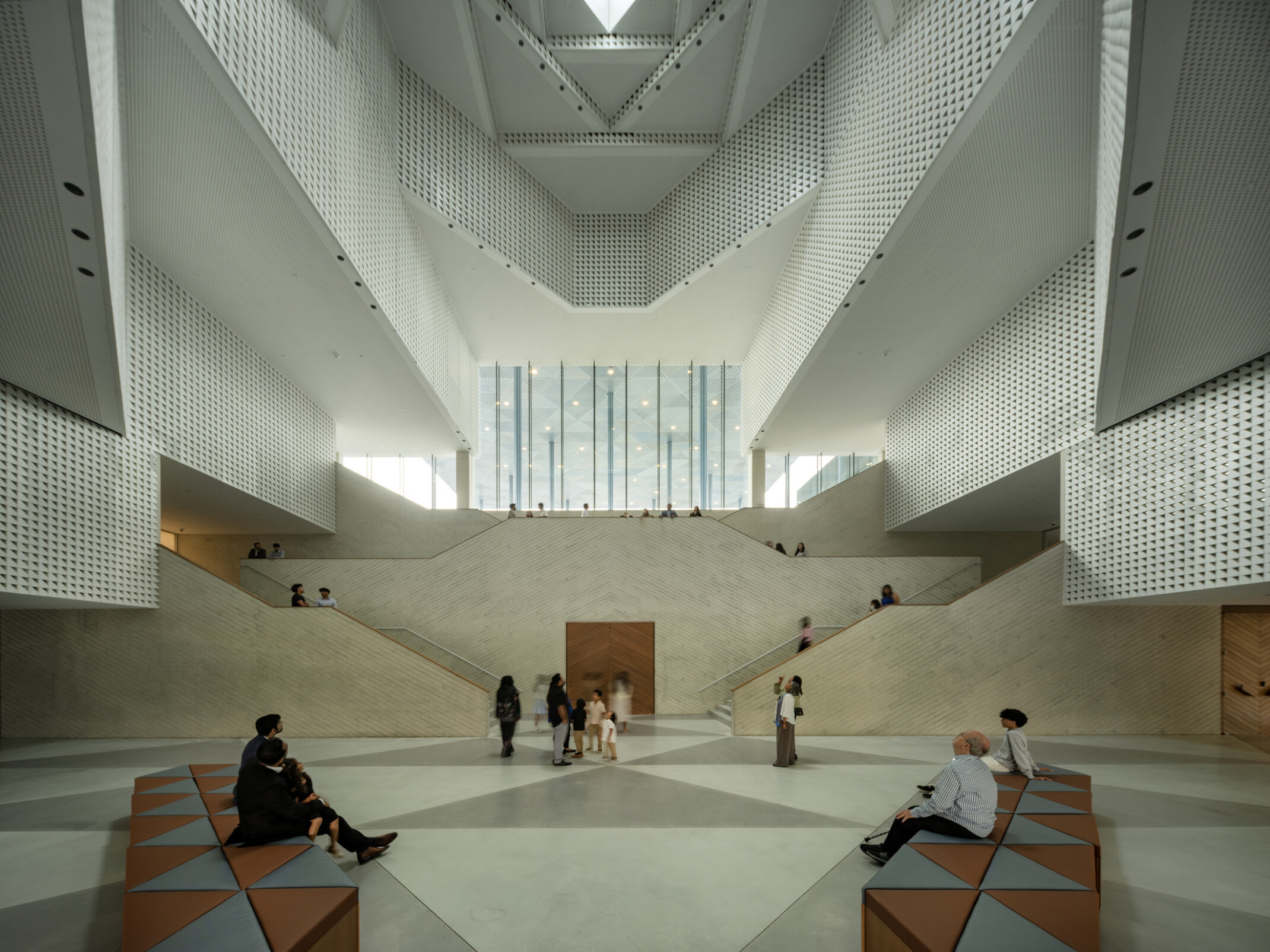 Atrium with perforated walls rising multiple stories around a dramatic angular staircase. Natural light floods through the oculus above and tall glass windows. Visitors move through the bright, open space, some seated in the foreground.