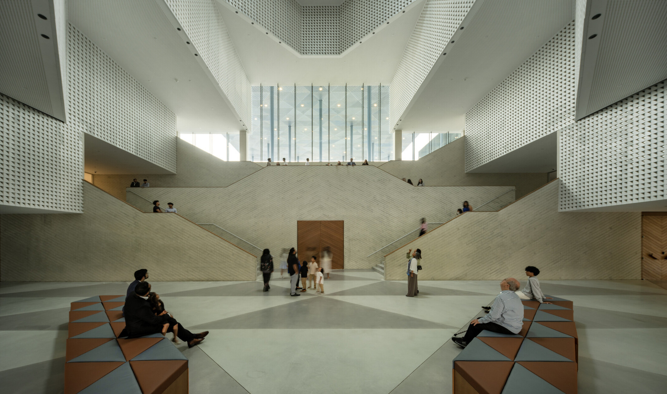 Atrium with perforated walls rising multiple stories around a dramatic angular staircase. Natural light floods through the oculus above and tall glass windows. Visitors move through the bright, open space, some seated in the foreground.