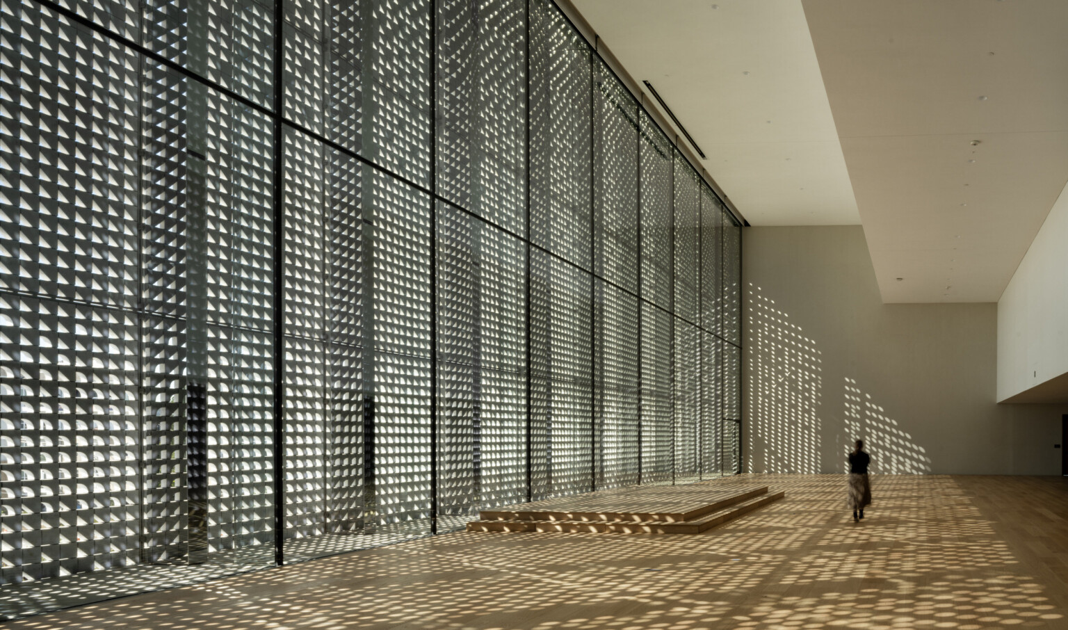 A spacious modern interior with floor-to-ceiling windows casting geometric shadows. A single person walks past a podium across warm wood floors.