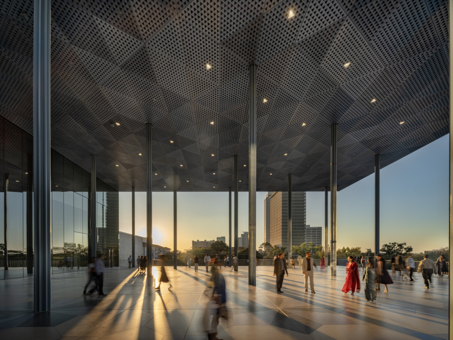 People walk under a large, modern pavilion with geometric patterned ceiling at sunset.