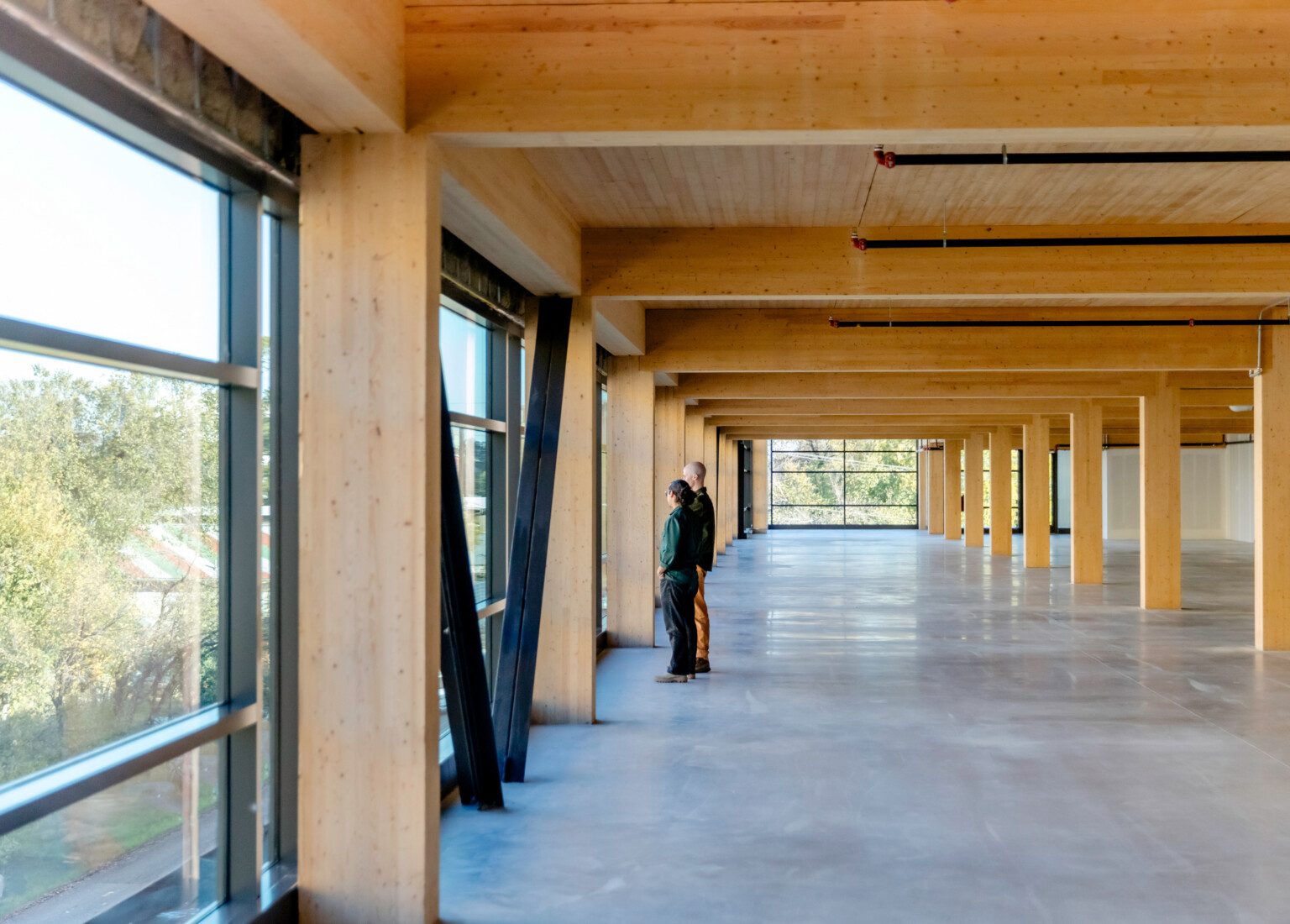 Large open space with windows on the left and back wall; two people looking out the left wall; Natural light wood accents the buildings beams and ceilings