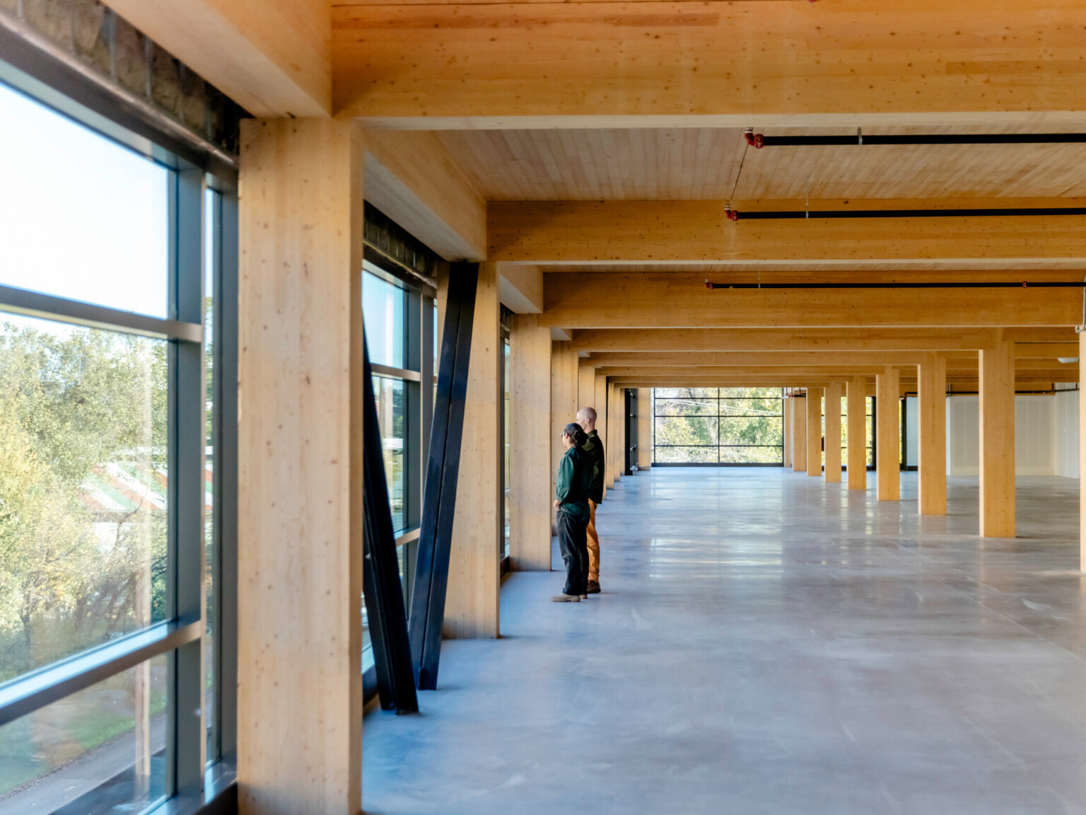 Large open space with windows on the left and back wall; two people looking out the left wall; Natural light wood accents the buildings beams and ceilings