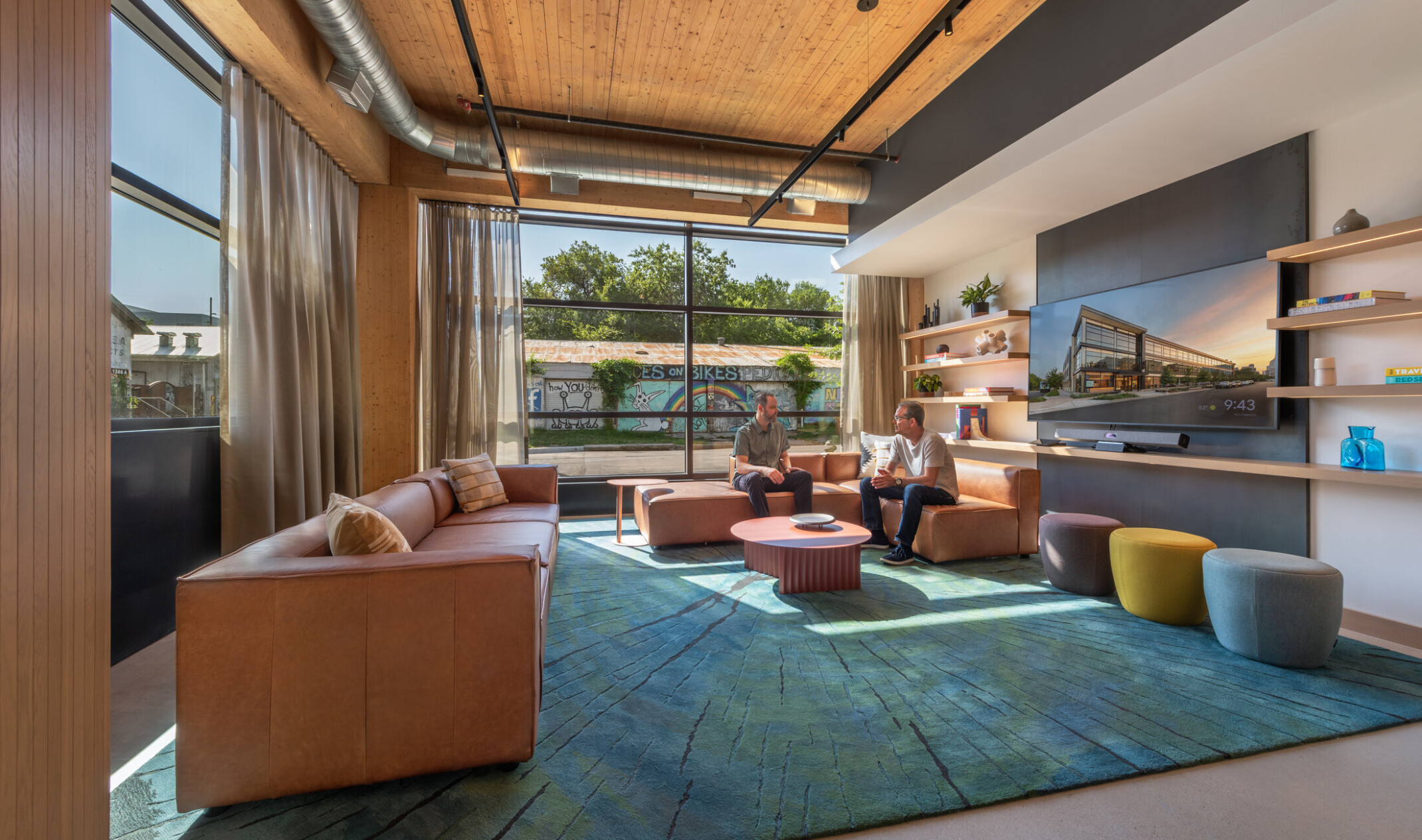 Modern seating area filled with brown leather couches, rug in shades of blue, ottomans, walls with shelves and TV and a colorful mural outside the windows