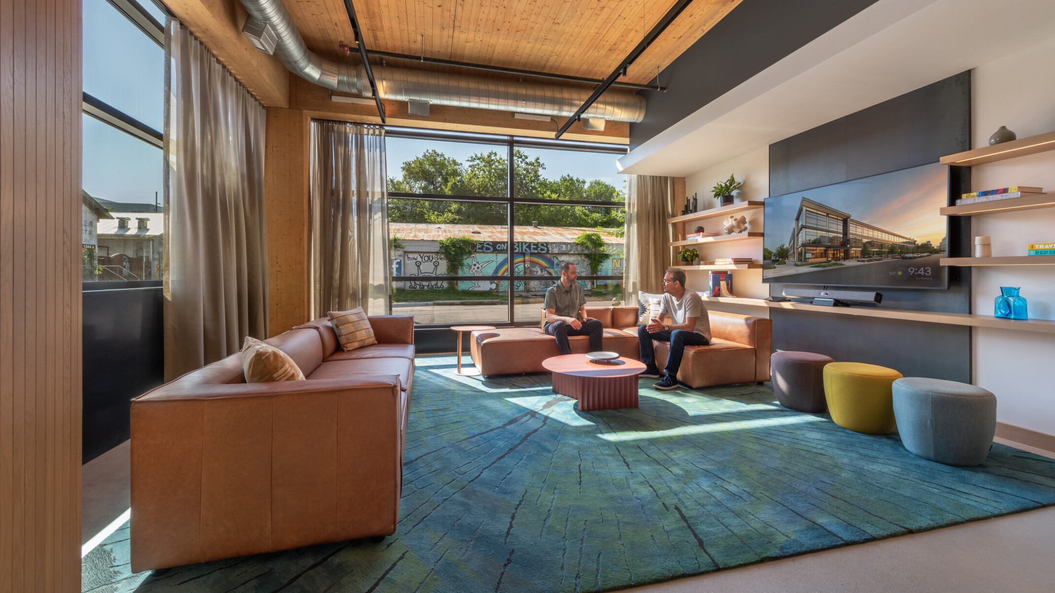 Modern seating area filled with brown leather couches, rug in shades of blue, ottomans, walls with shelves and TV and a colorful mural outside the windows