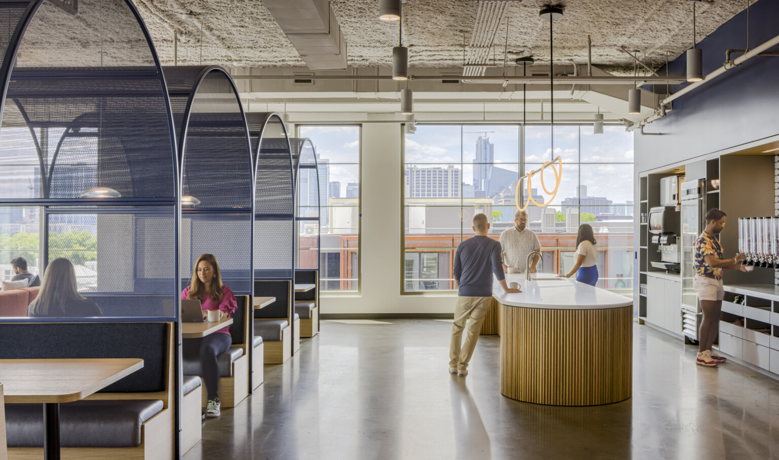 A modern office with booth seating, open spaces, and city views. Employees collaborate near a counter, with an industrial-style ceiling above.