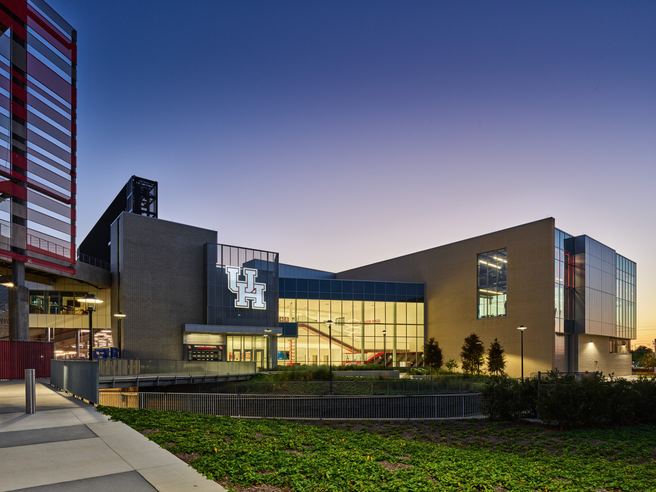 A modern building with large glass windows and an illuminated “UH” logo shown at dusk, with landscaped greenery in the foreground and a gradient evening sky behind it.
