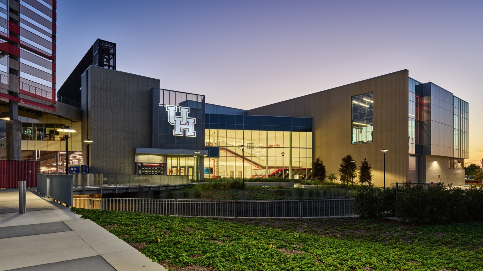 A modern building with large glass windows and an illuminated “UH” logo shown at dusk, with landscaped greenery in the foreground and a gradient evening sky behind it.