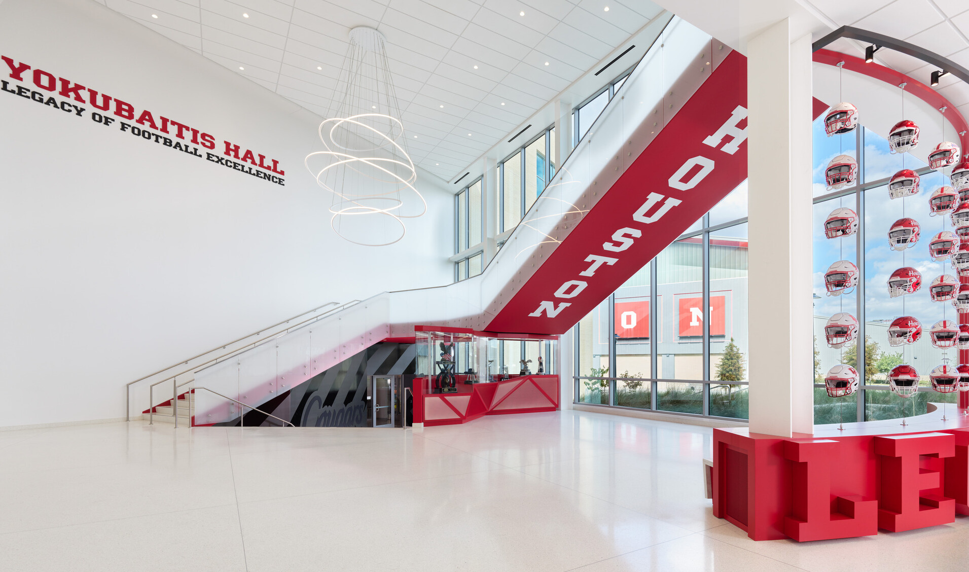 A bright, modern lobby with white walls and large windows, featuring a sweeping staircase with red accents, a wall displaying “Yokubaitis Hall Legacy of Football Excellence,” and a curved glass case showcasing rows of red football helmets.