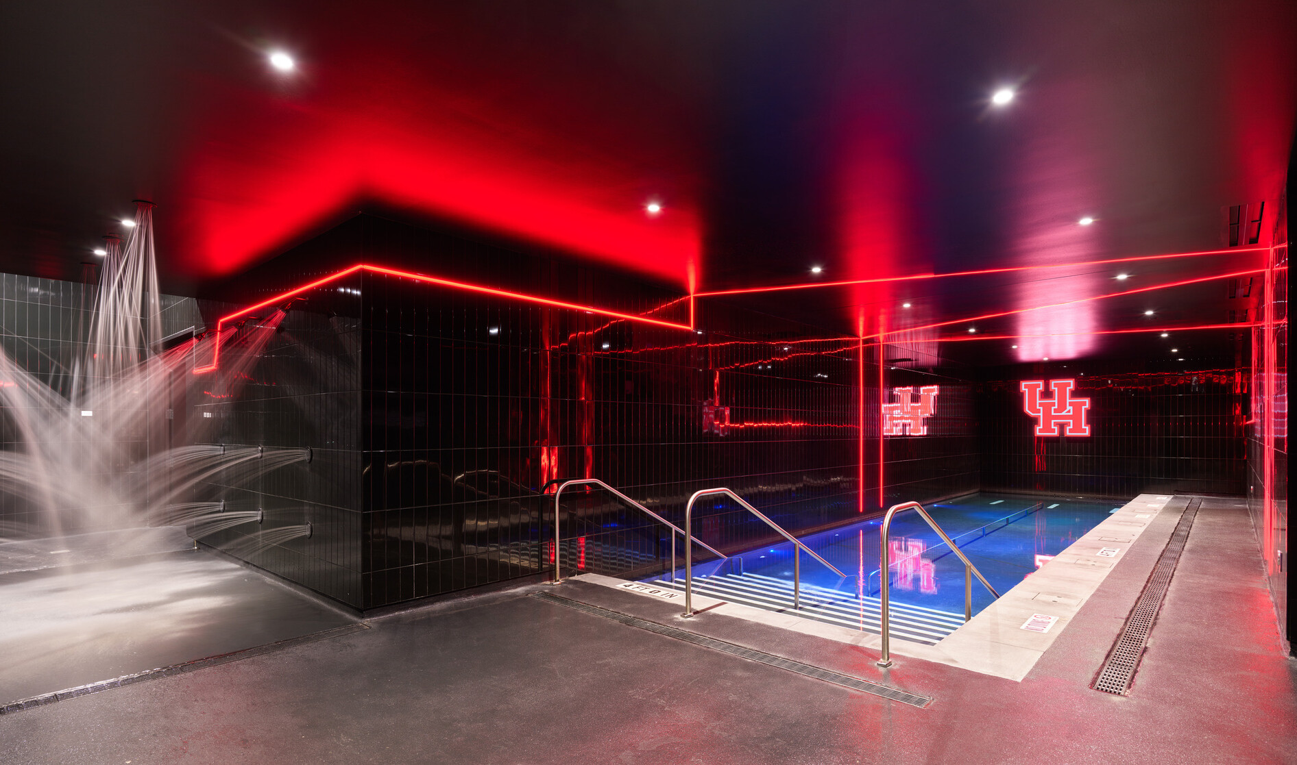 A dark hydrotherapy room featuring a lit soaking pool with red accent lighting, black tile walls, ceiling spotlights, and a separate area with cascading water jets on the left.