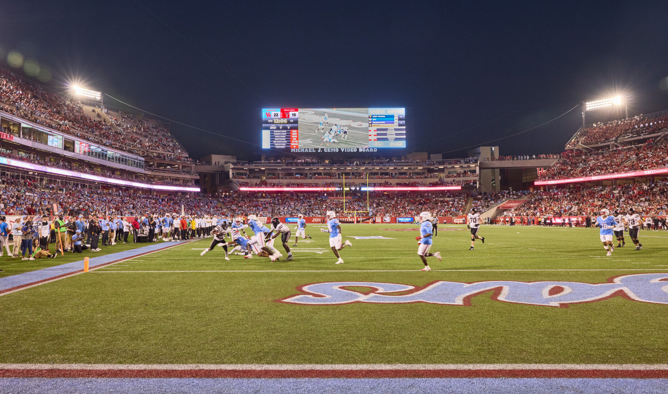 A nighttime football game in a packed stadium, showing players on the field near the end zone with a large illuminated scoreboard and crowd-filled stands in the background.