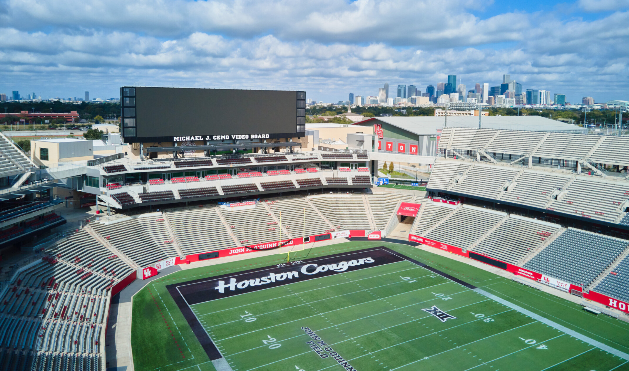 large outdoor football stadium with gray seating surrounding a green field marked “Houston Cougars,” featuring a massive video board on one end and a city skyline visible in the background under a bright, partly cloudy sky.