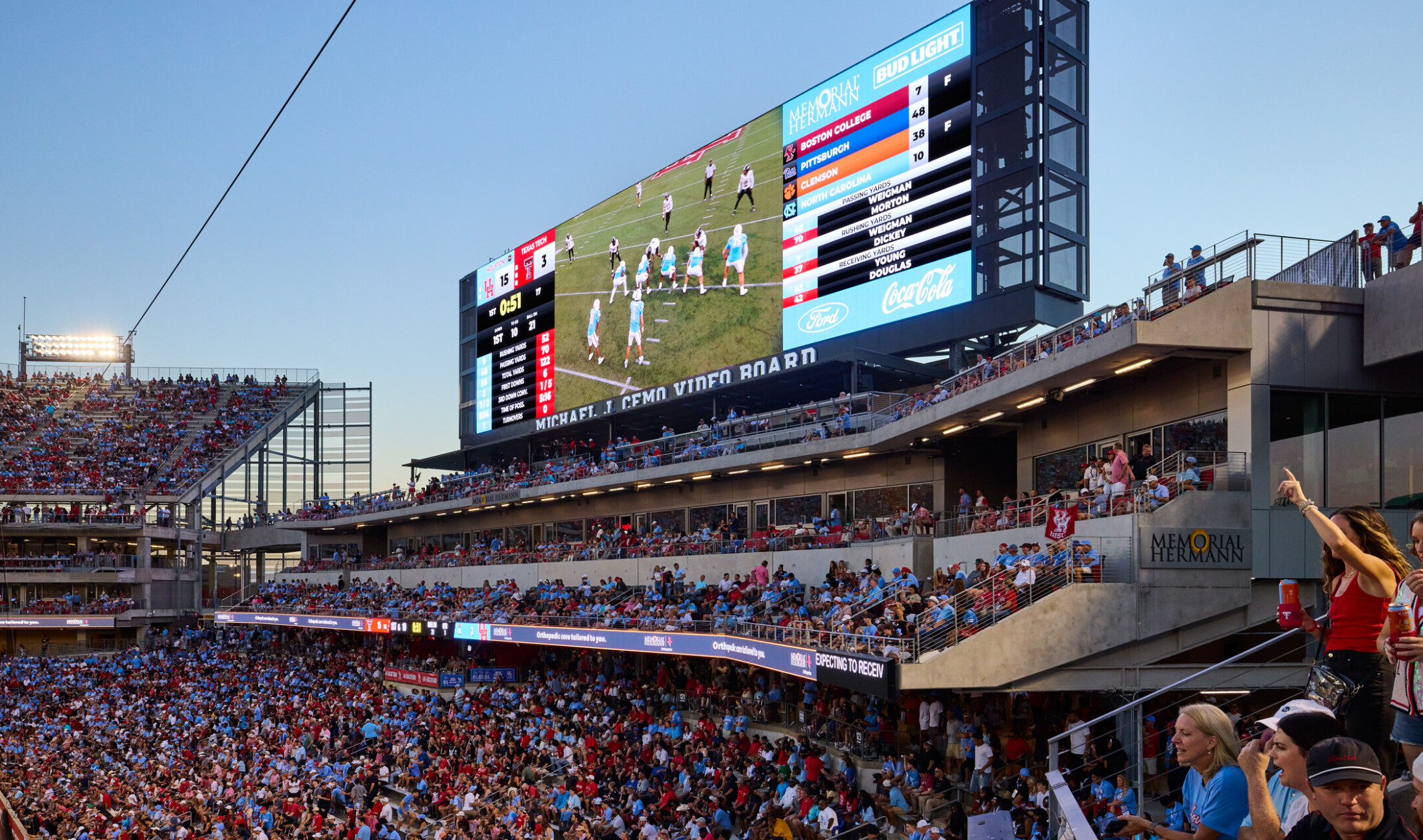 A large outdoor stadium filled with spectators, with multiple seating levels, an illuminated scoreboard displaying a football game, and fans standing and cheering along the concourse in the foreground.