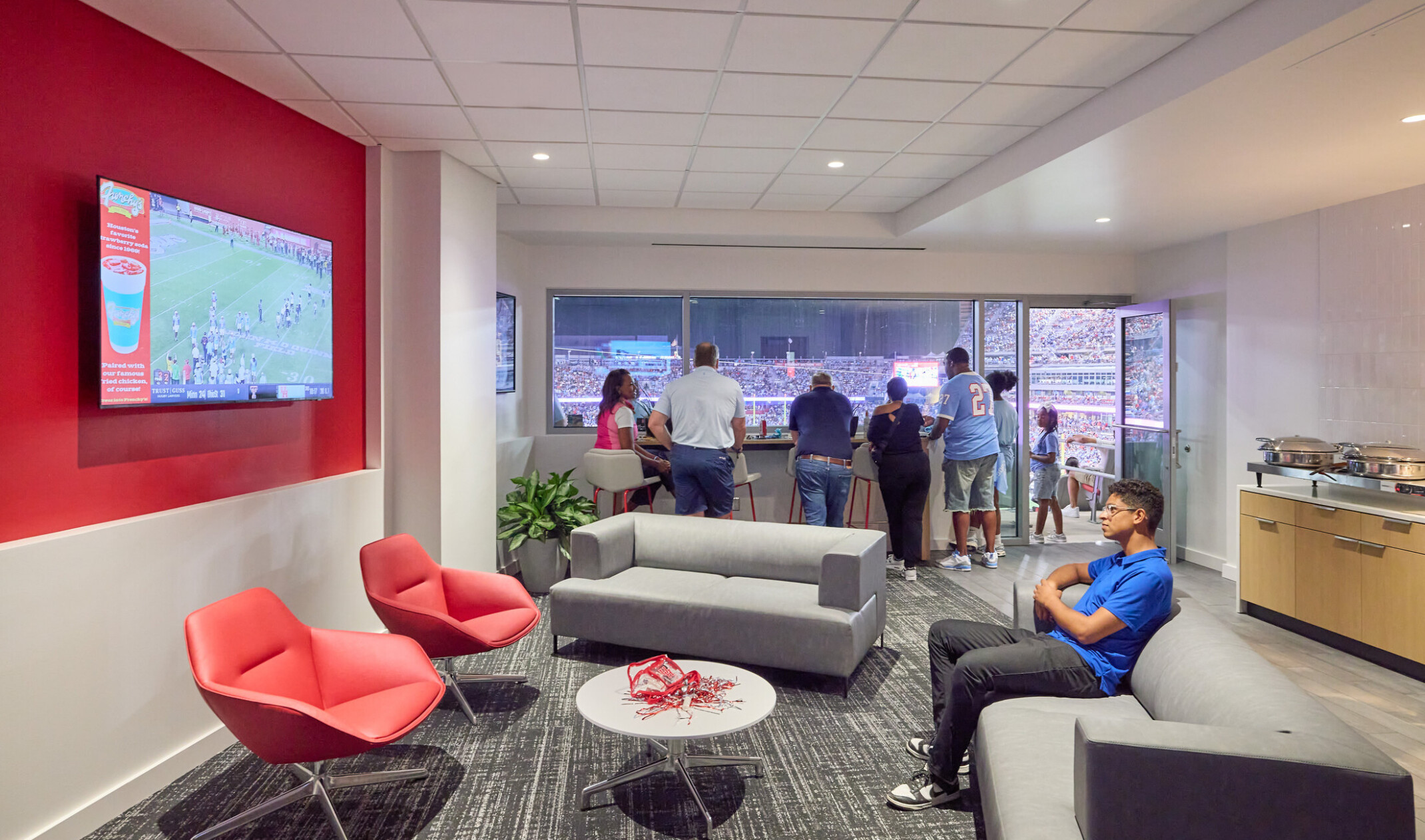 A lounge suite overlooking a stadium, featuring gray sofas, red lounge chairs, a wall mounted TV showing a football game, and a group of people standing at a counter by large windows with a view of the field.
