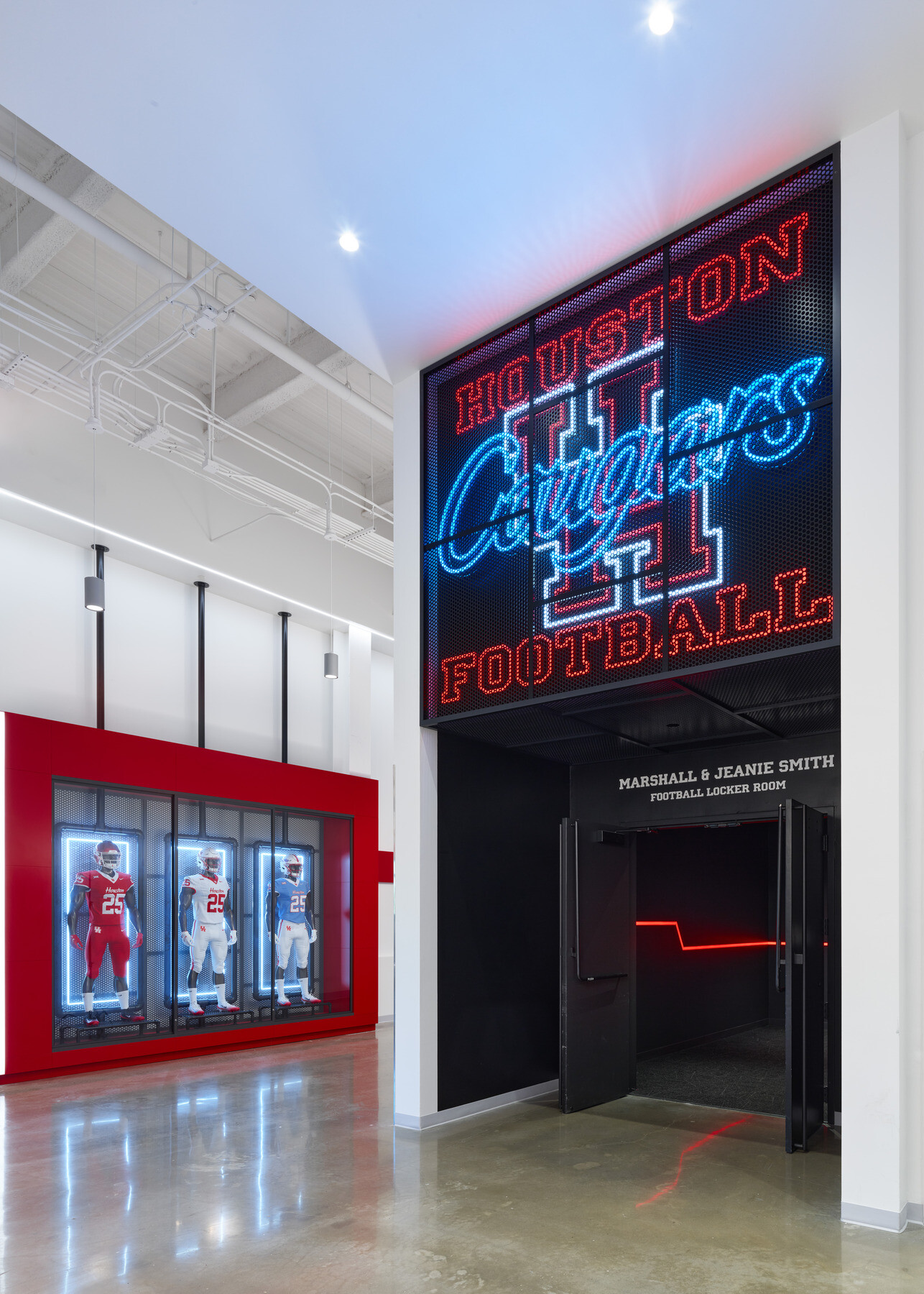 A bright lobby area with a large illuminated “Houston Cougars Football” sign above the entrance to a locker room, and a display of mannequin football uniforms in a red-framed case along the adjacent wall.