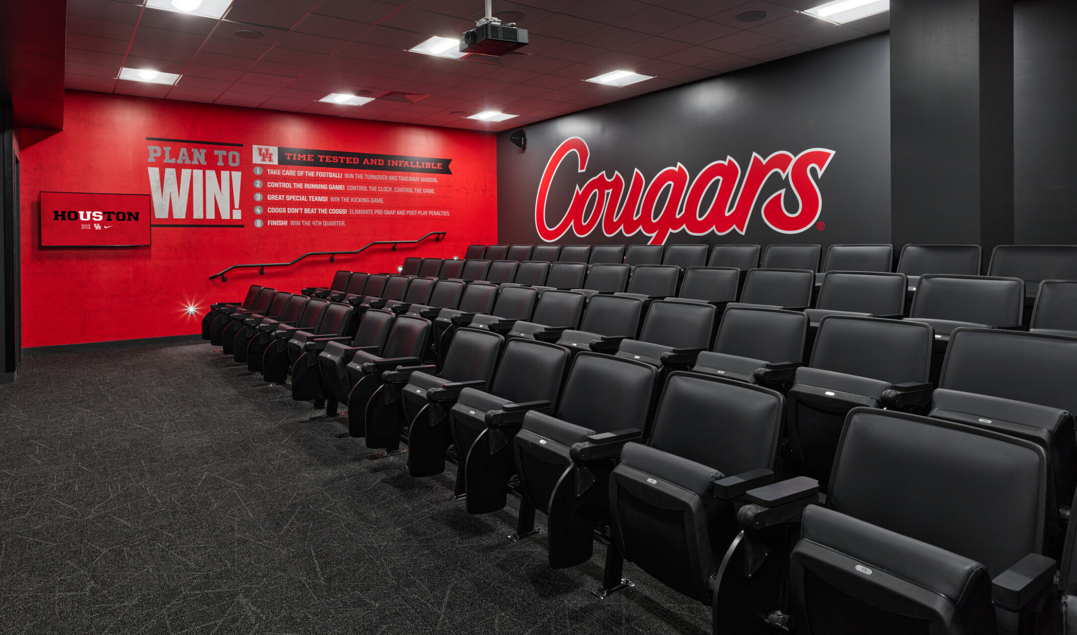 A tiered team meeting room with rows of black theater-style seats facing a red accent wall with motivational text and a large “Cougars” logo on the side wall.