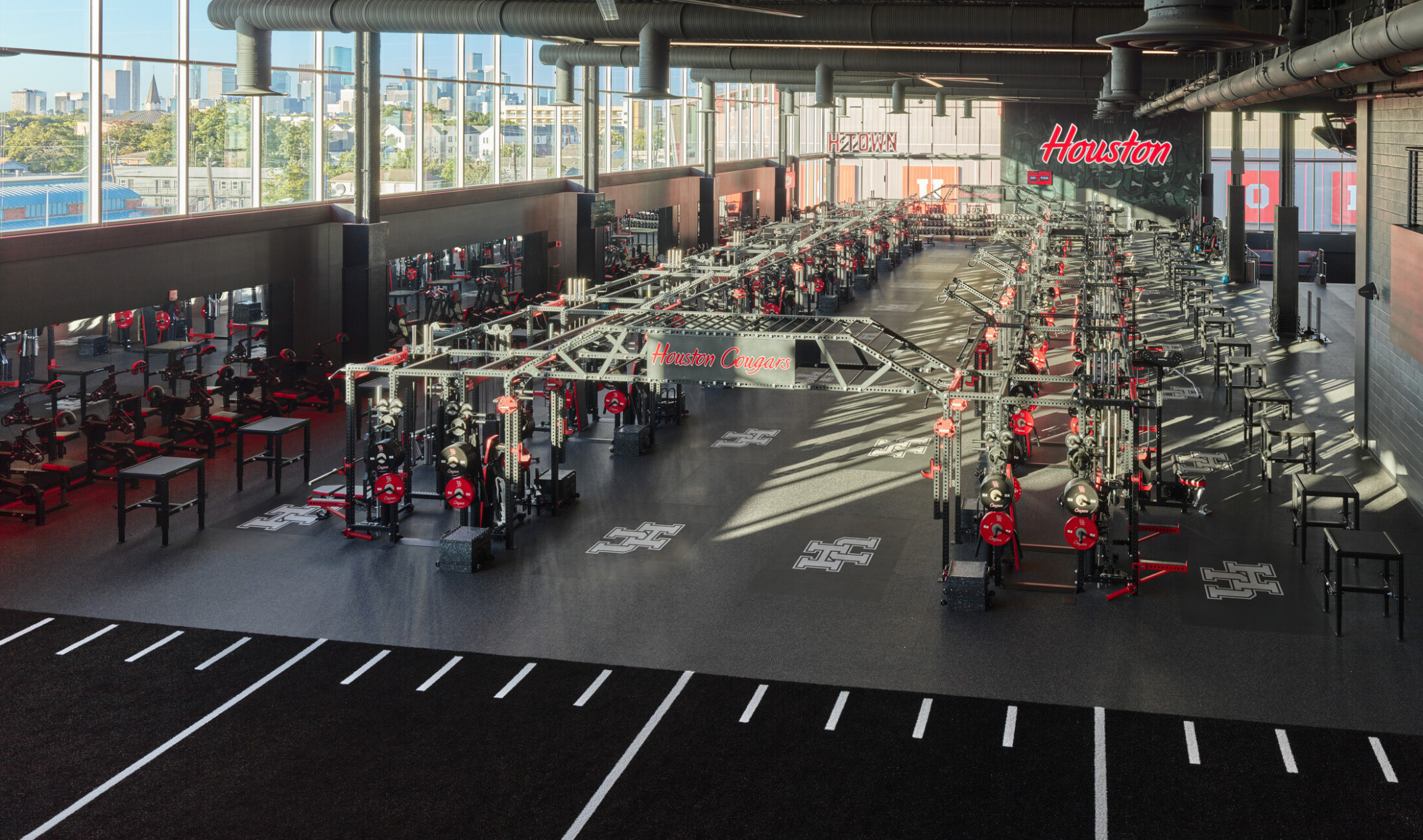A large, open training facility filled with weightlifting racks and equipment arranged in rows, surrounded by floor‑to‑ceiling windows with views of the city skyline