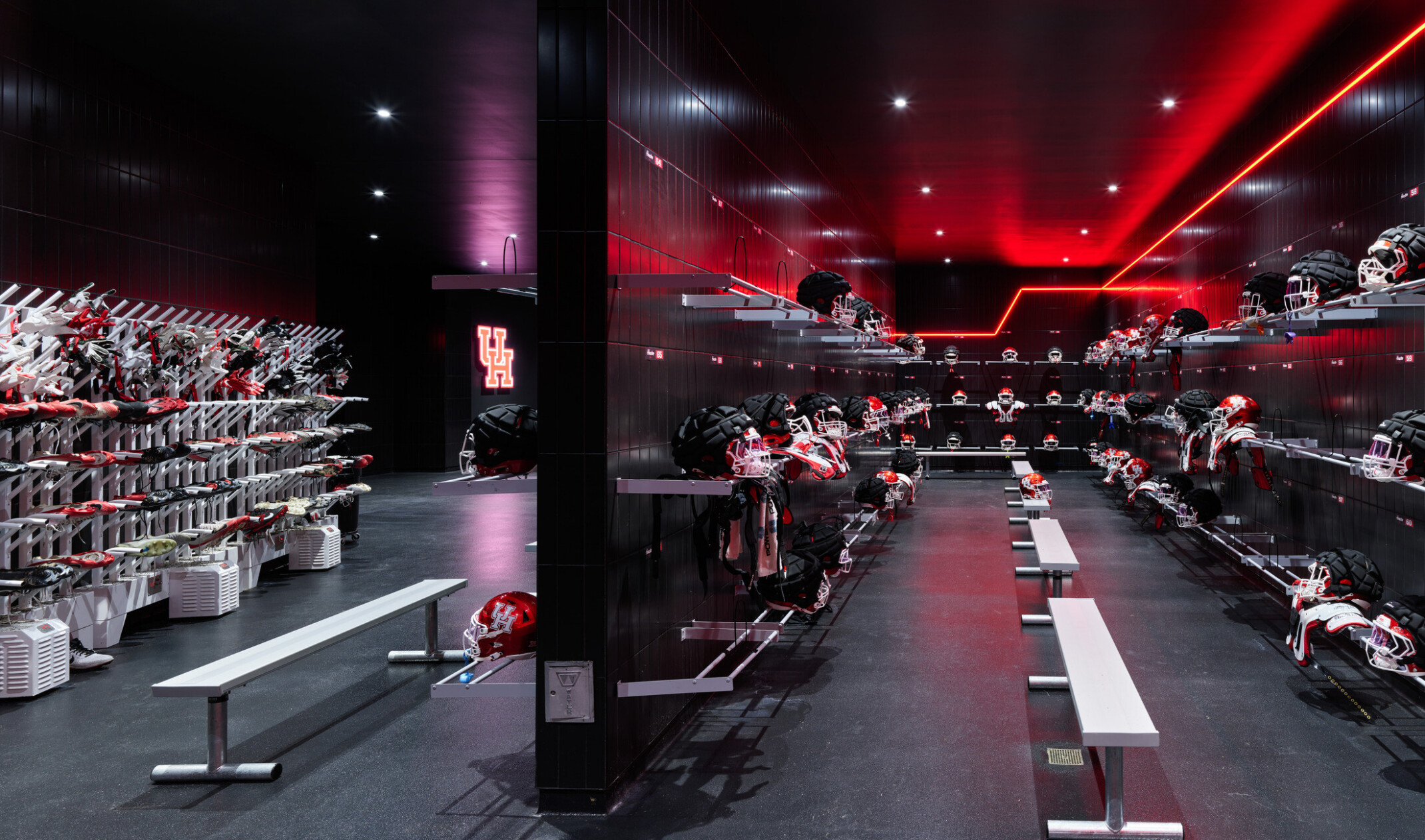 A dark equipment room lined with shelves of football helmets and pads, illuminated by red accent lighting, with metal benches positioned along the center walkway.
