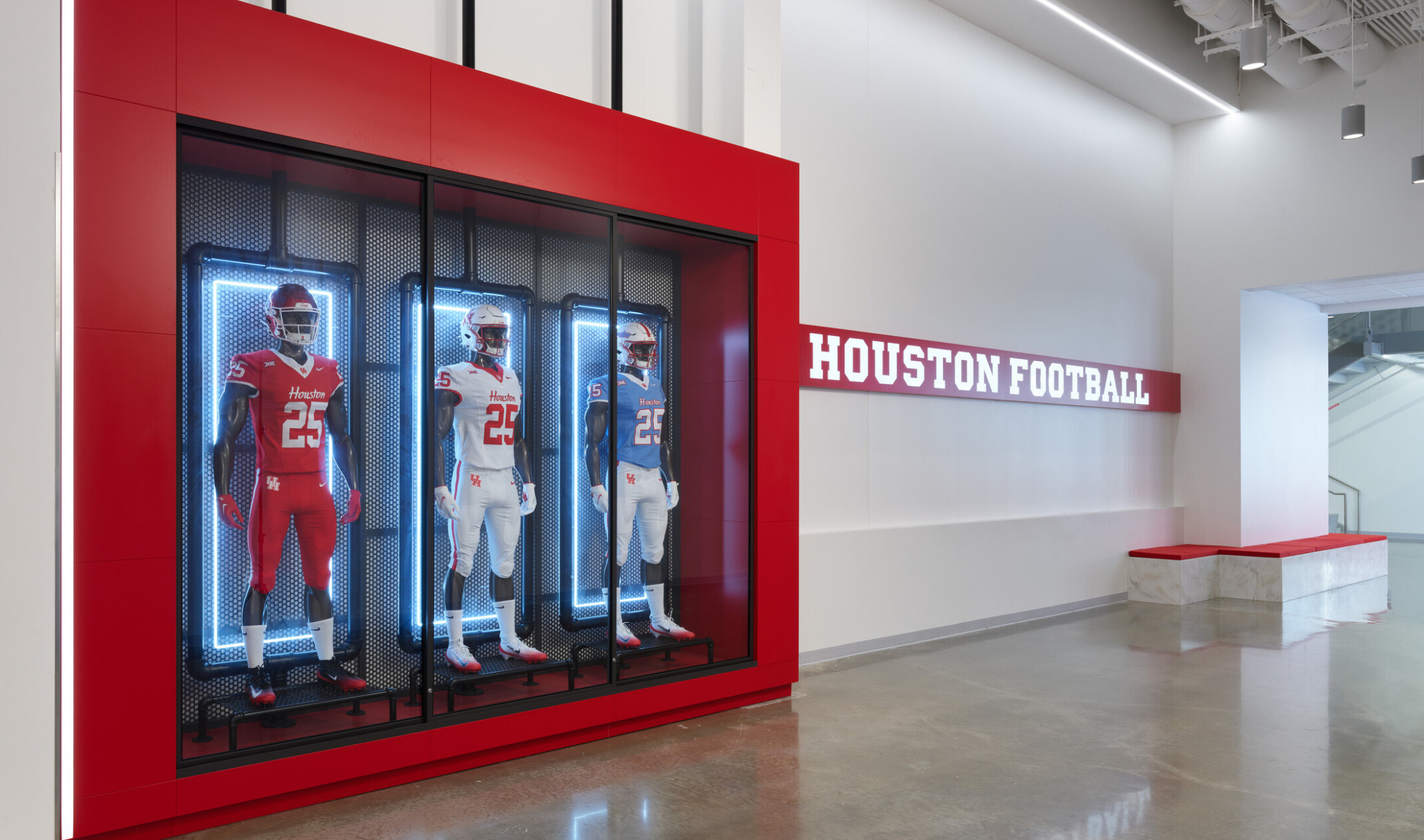A bright lobby hallway featuring a large red-framed display case with three illuminated mannequin football uniforms and a wall sign reading “Houston Football,” with polished concrete floors and exposed ceiling fixtures.