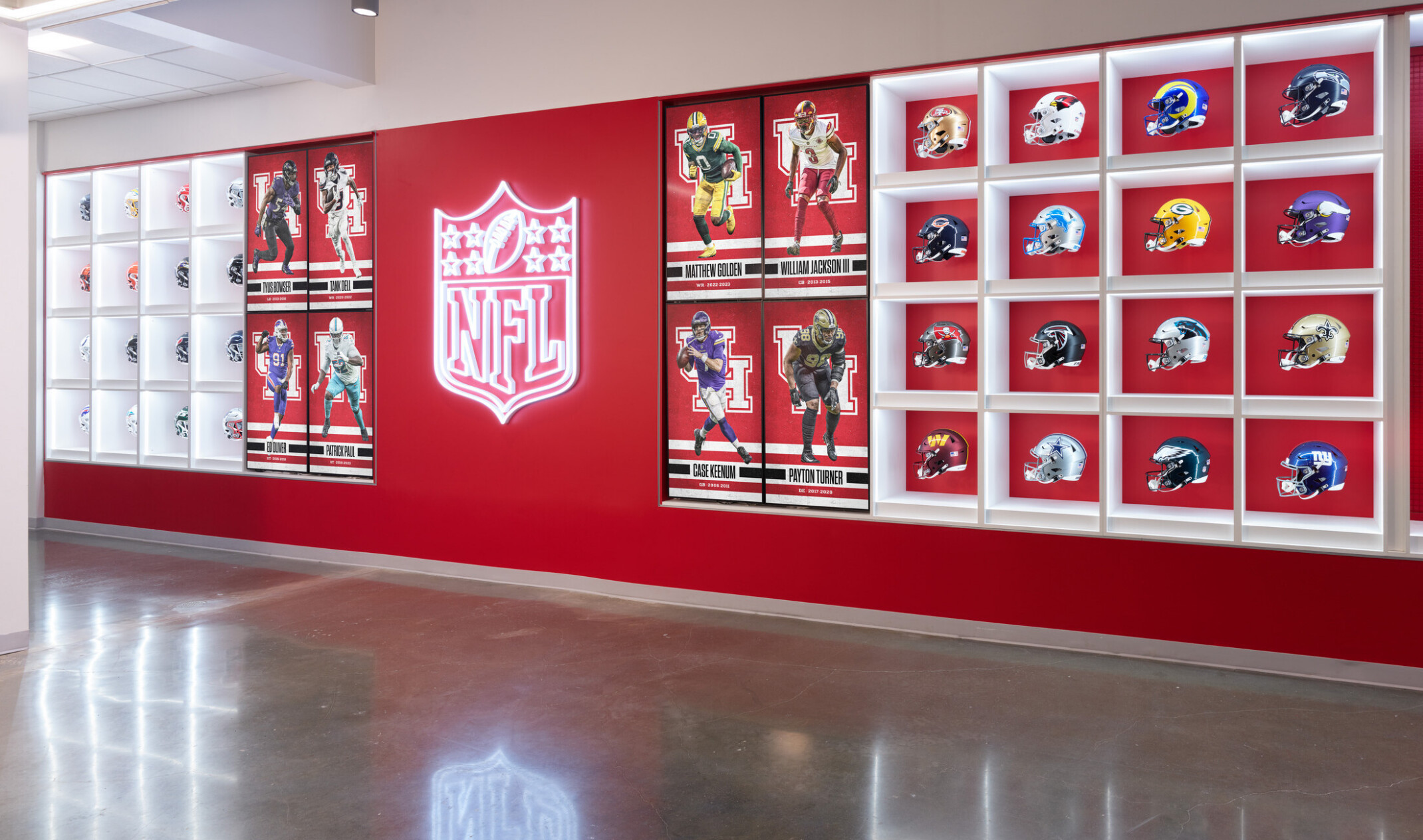 A bright hallway display featuring a large illuminated NFL logo flanked by framed images of football players and a grid of shelves showcasing various team helmets against a red and white backdrop.
