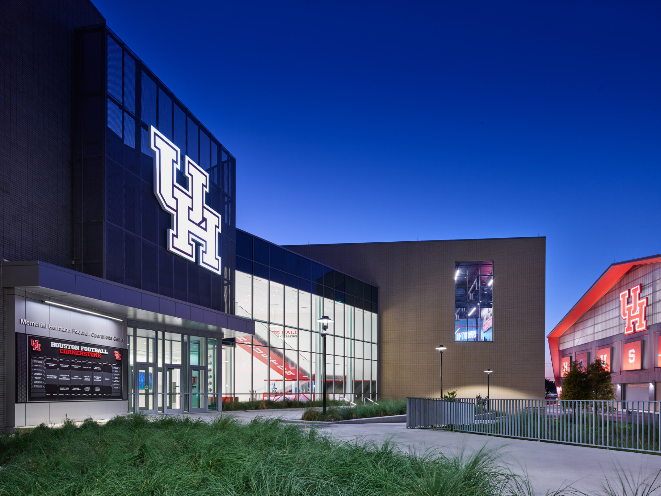 Exterior view of the University of Houston football operations facility at dusk, featuring a large illuminated “UH” logo on a glass façade, modern architecture, landscaped walkways, and a neighboring athletic building with a glowing red roofline and University of Houston branding