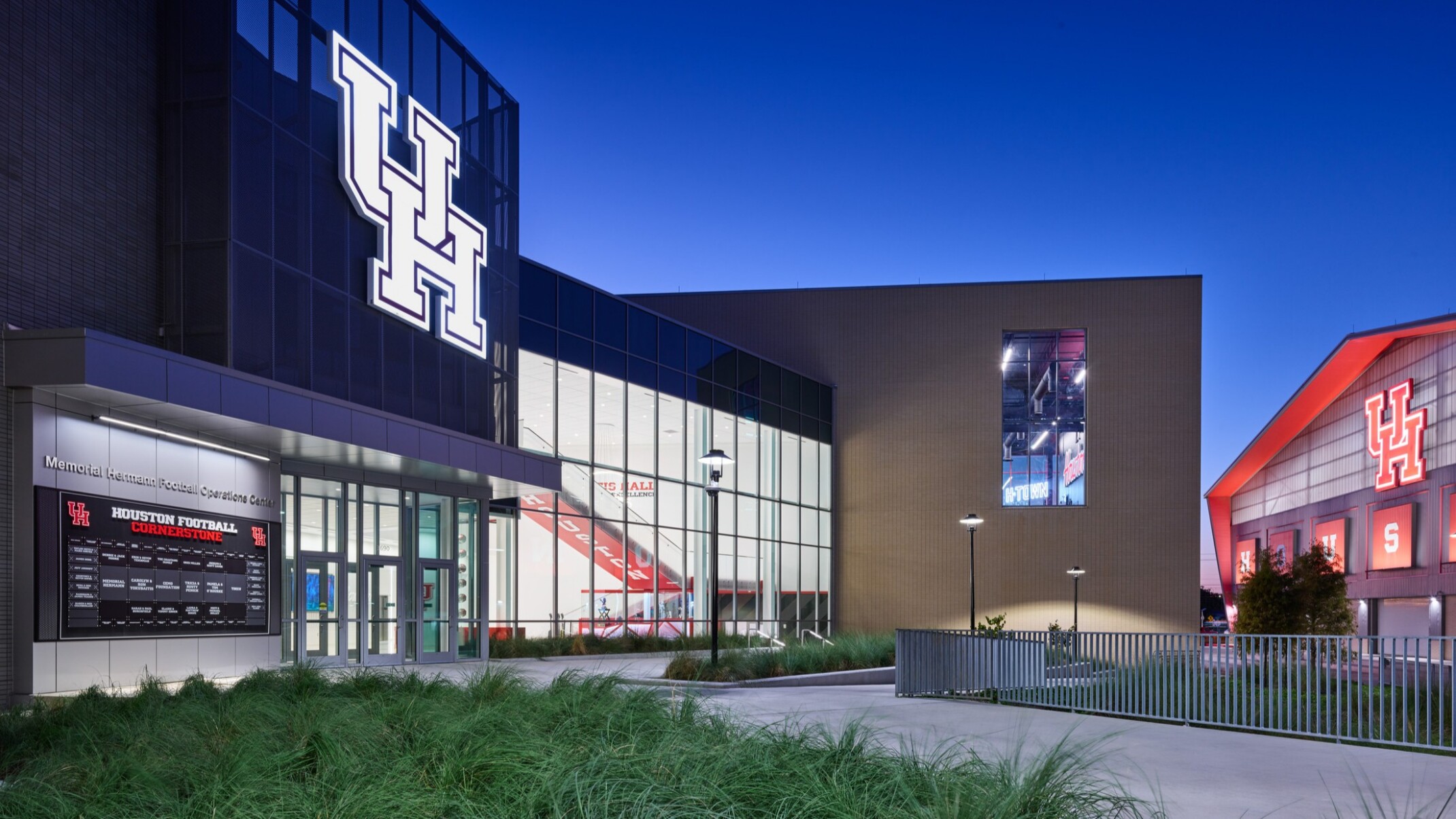 Exterior view of the University of Houston football operations facility at dusk, featuring a large illuminated “UH” logo on a glass façade, modern architecture, landscaped walkways, and a neighboring athletic building with a glowing red roofline and University of Houston branding