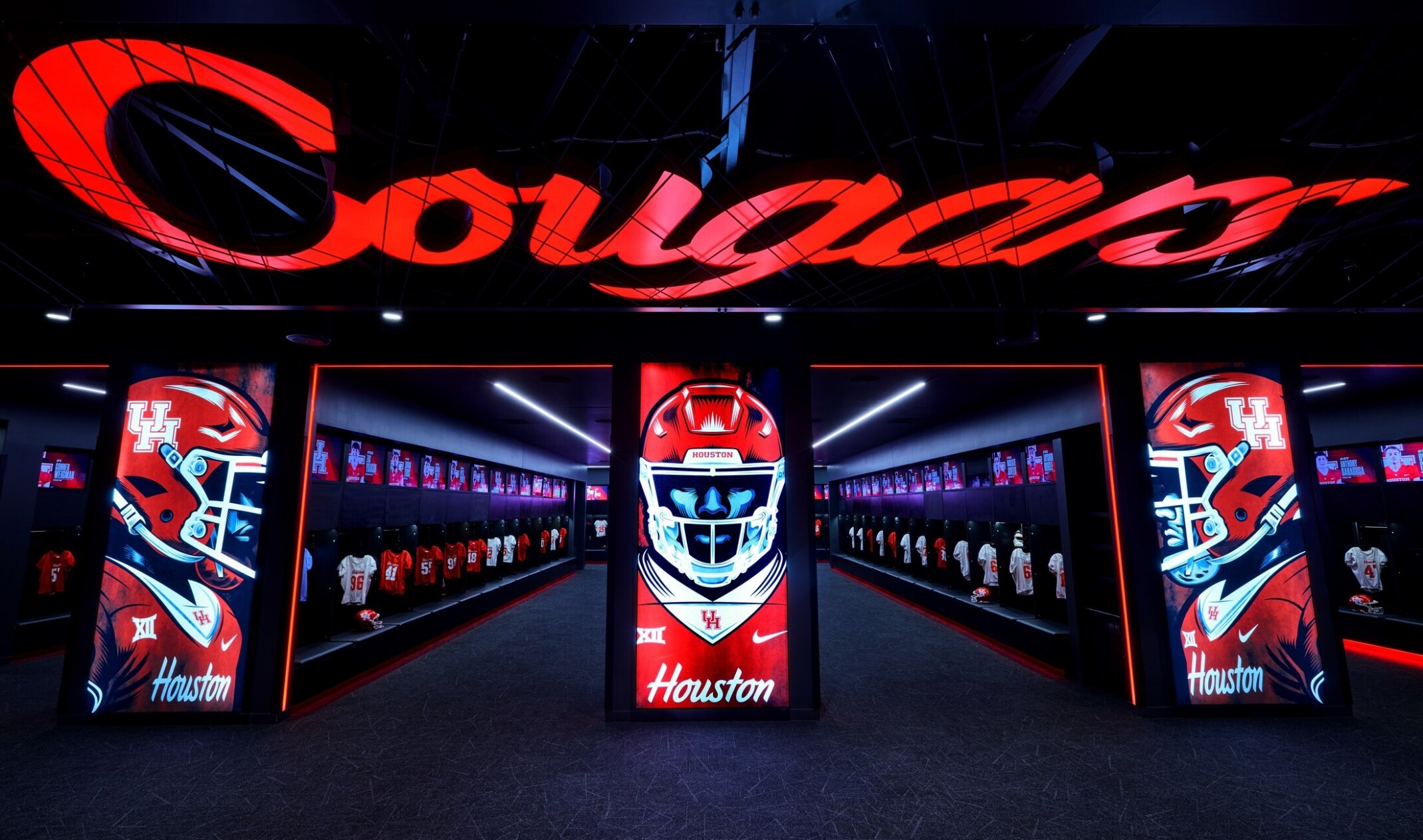 A dimly lit athletic locker room entrance featuring large illuminated displays of football helmets and jerseys, with rows of red and white team lockers visible on both sides and a glowing “Cougars” sign overhead