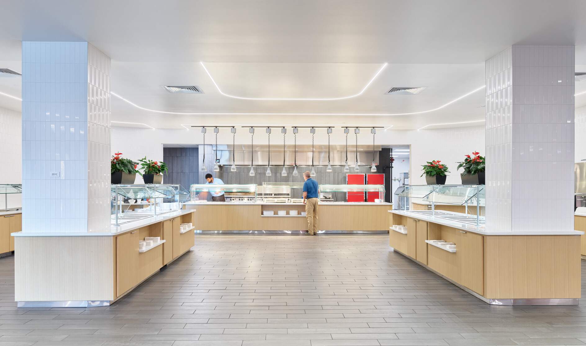 A bright, modern cafeteria serving area with light wood counters, glass food display stations, white tile columns, and overhead heat lamps, with two people working behind the central counter and potted plants placed throughout the space