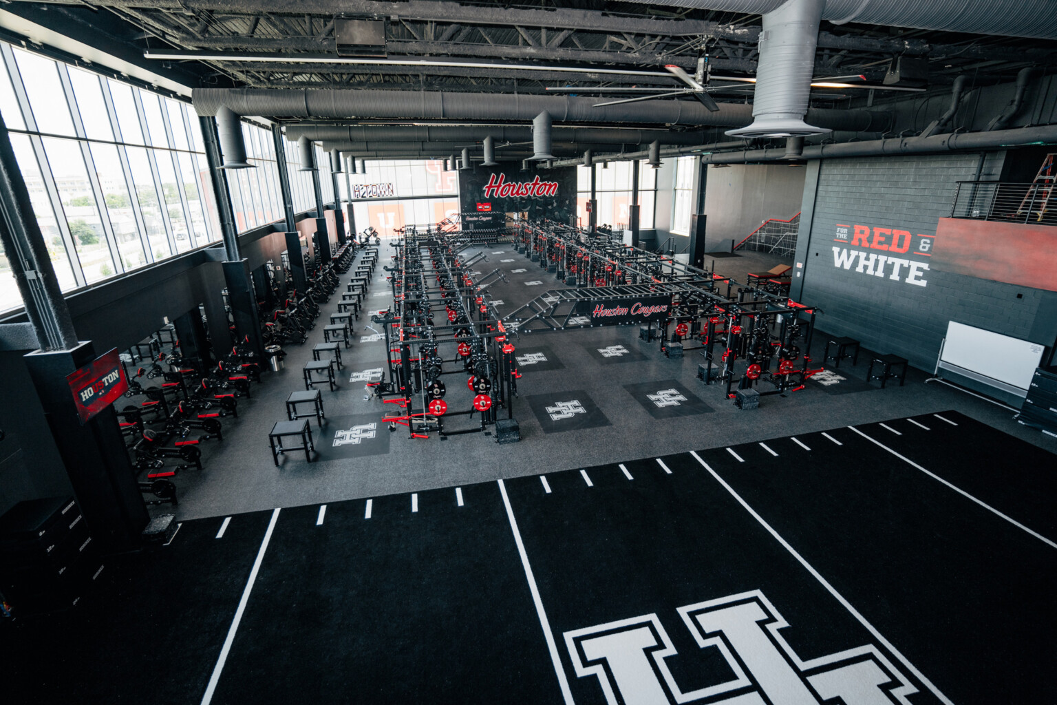University of Houston football operations center weightroom with large UH logos on a indoor field space in front of 2 rows of weight lifting machinery