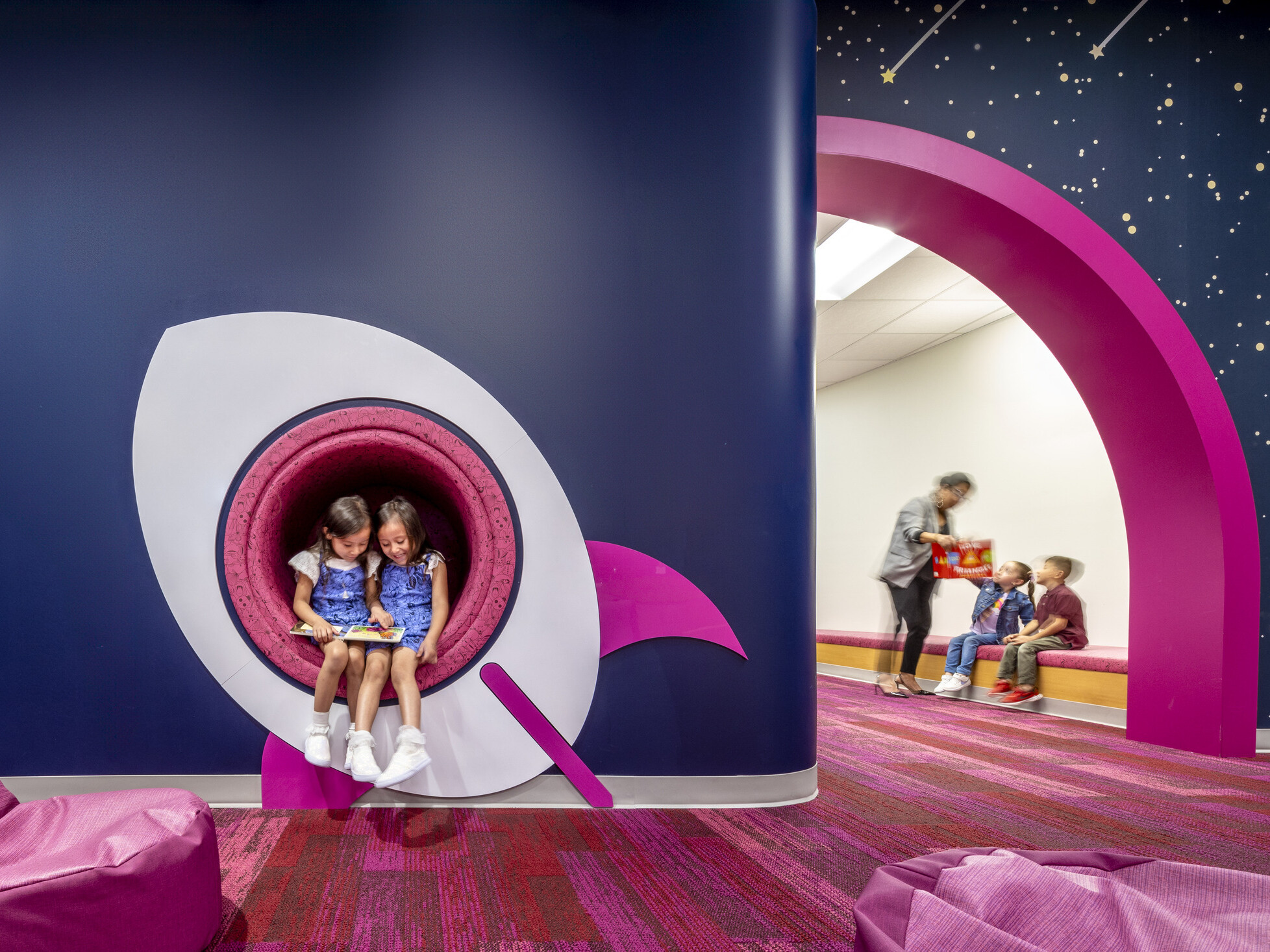 kids reading a book in a cutout window of a space ship mural. Blue walls with pink accents at a early excellence academy.