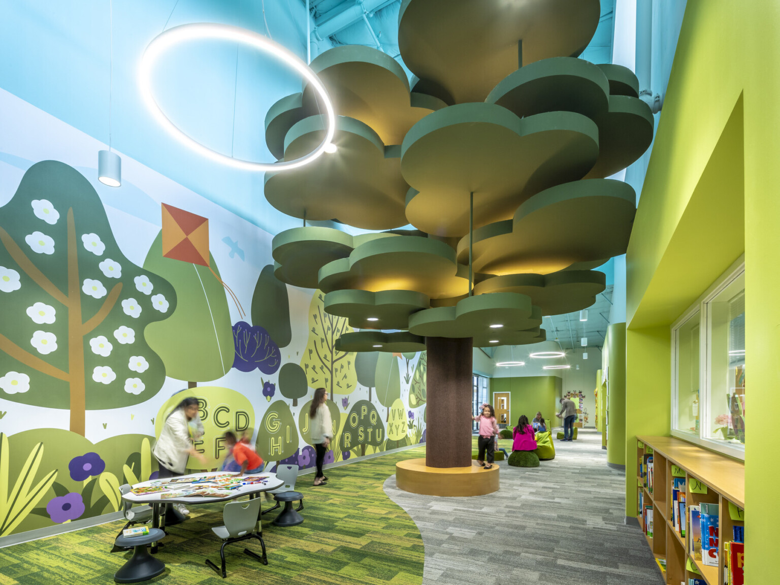 interior of a k-12 school designed to look like the outdoors; a large scultpural tree sits in the middle of the room while wall finishes, paint colors, and layout mimic the forest