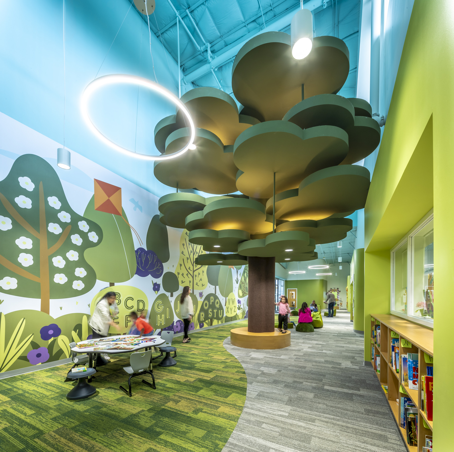 interior of a k-12 school designed to look like the outdoors; a large scultpural tree sits in the middle of the room while wall finishes, paint colors, and layout mimic the forest