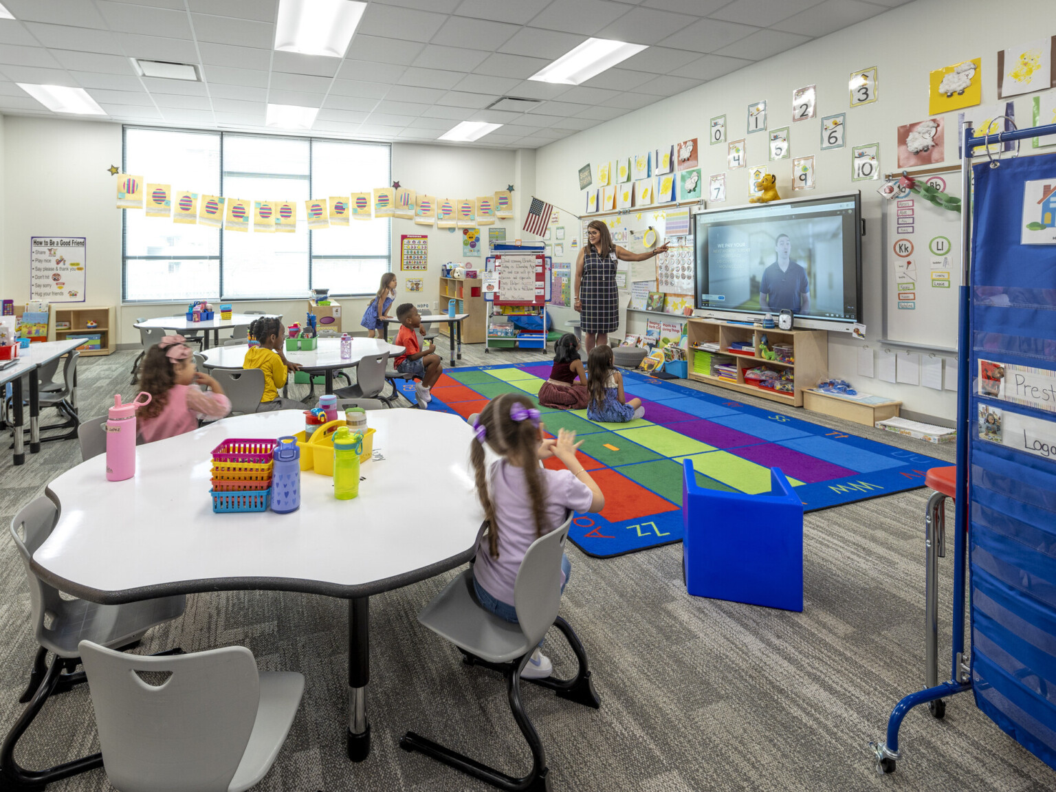 Classroom at an early excellence academy with colorful rug on top of grey floors with tables filled with students and a teacher standing in front of a monitor hanging on the wall