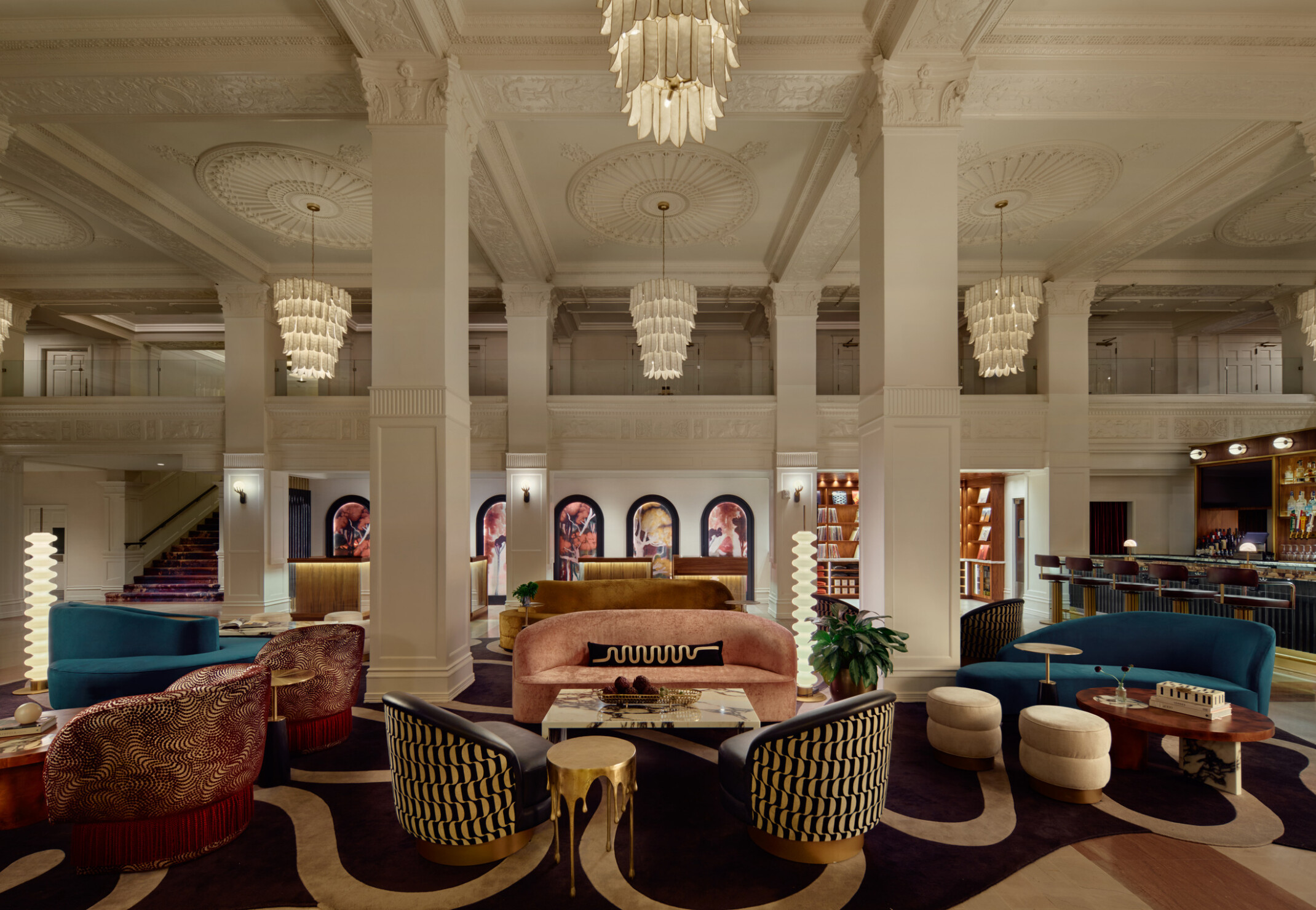 Gunter Hotel lobby lounge with jewel-toned seating, original columns and rosette ceiling, softly glowing chandeliers overhead, check-in desk in the background, and the Keystone Club bar and vinyl library to the right.