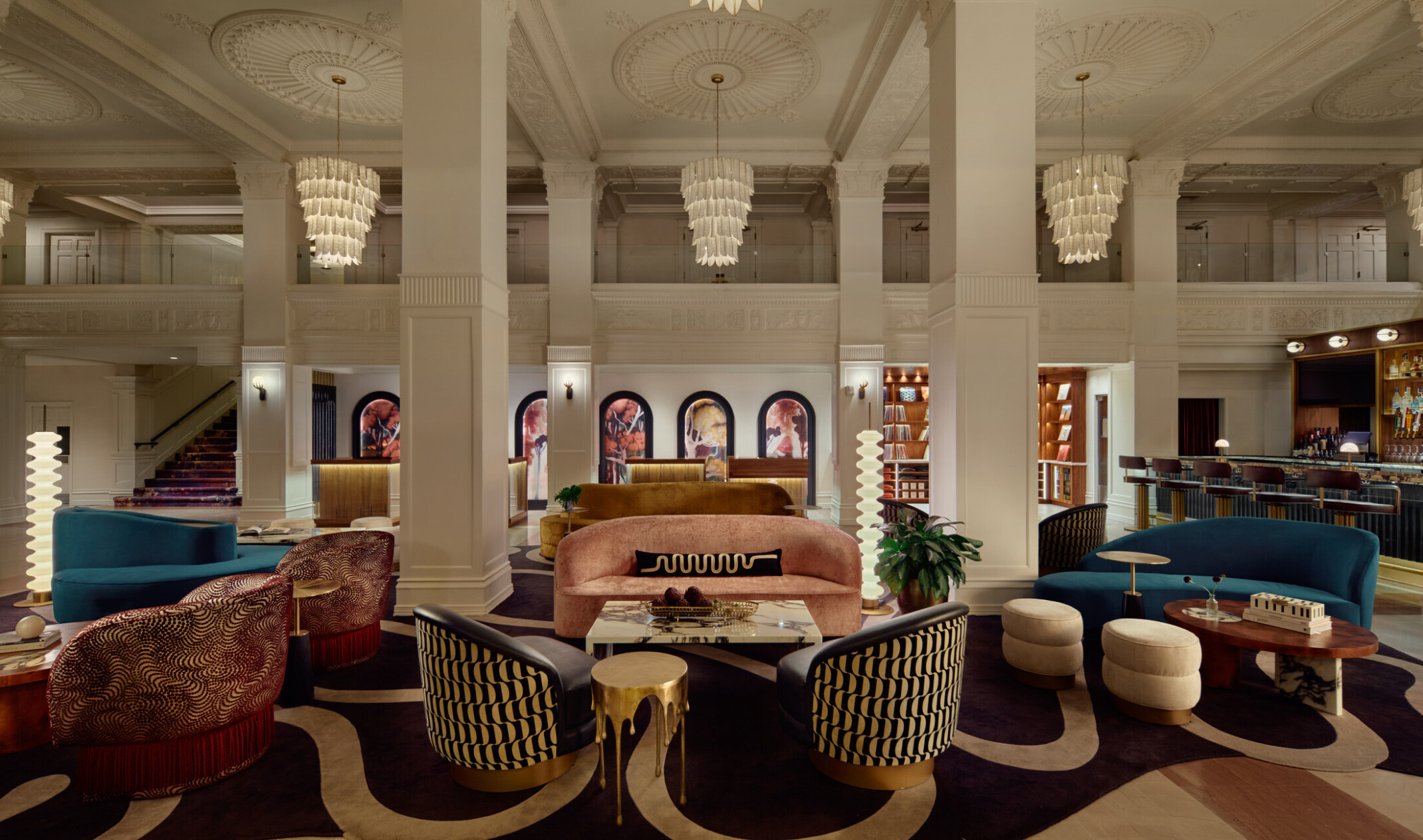 Gunter Hotel lobby lounge with jewel-toned seating, original columns and rosette ceiling, softly glowing chandeliers overhead, check-in desk in the background, and the Keystone Club bar and vinyl library to the right.