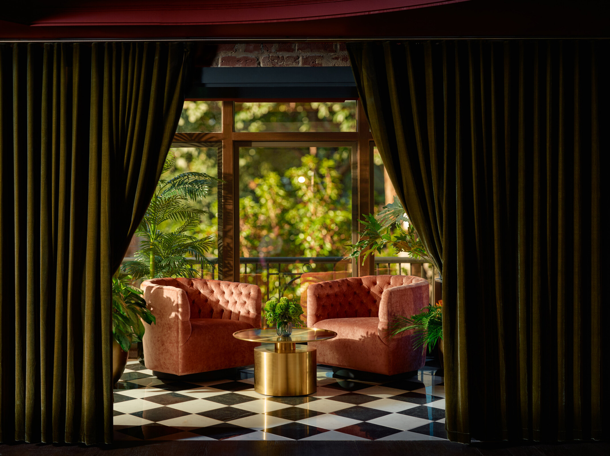 Green velvet drapes part to reveal the Studio lounge’s glass-domed terrace, where two blush pink chairs sit among lush potted plants with a view of street trees.