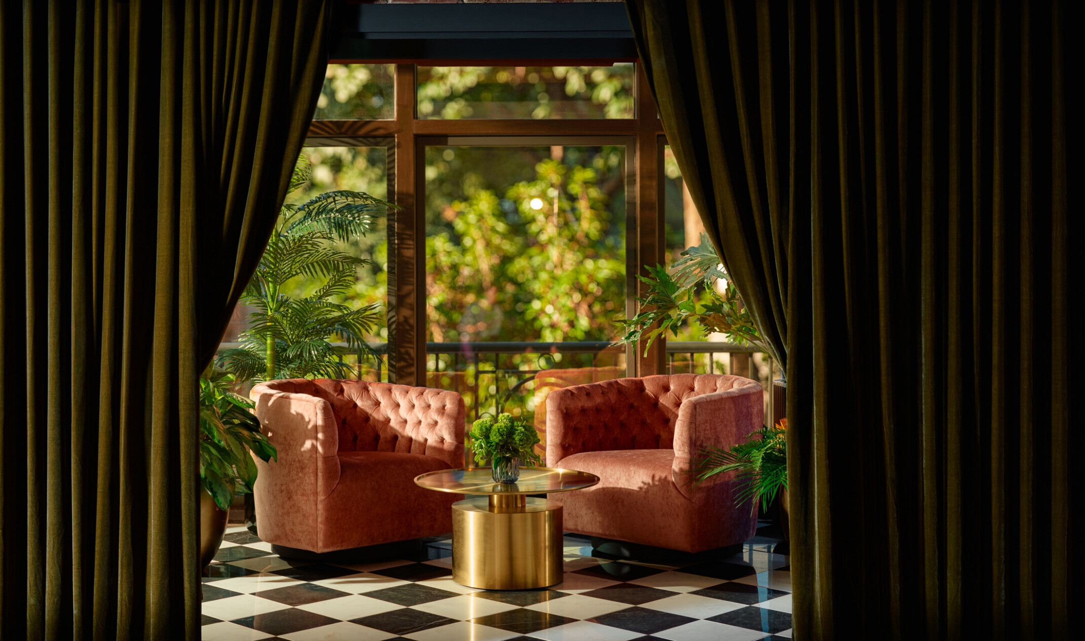 Green velvet drapes part to reveal the Studio lounge’s glass-domed terrace, where two blush pink chairs sit among lush potted plants with a view of street trees.