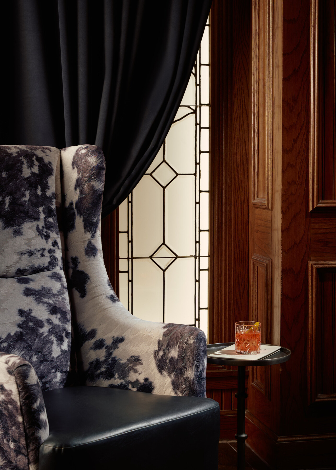 Detail of Bar 414 at the Gunter Hotel, where a glowing faux window partly draped in blue velvet frames a cowhide chair, wood paneling, and a waiting drink.