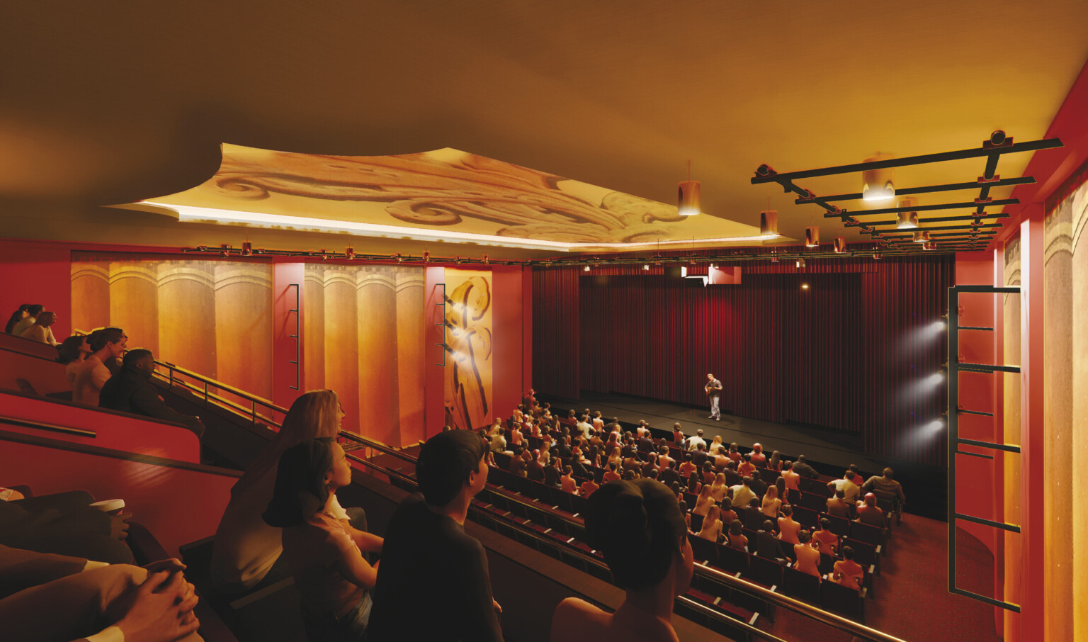View from an upper balcony inside a large auditorium, looking toward a stage where a person is speaking. The audience fills the lower seating area, and several people are visible in the balcony foreground. The space features warm amber and red tones, tall golden wall panels, and an ornate ceiling design. Dark curtains frame the stage, creating an elegant atmosphere