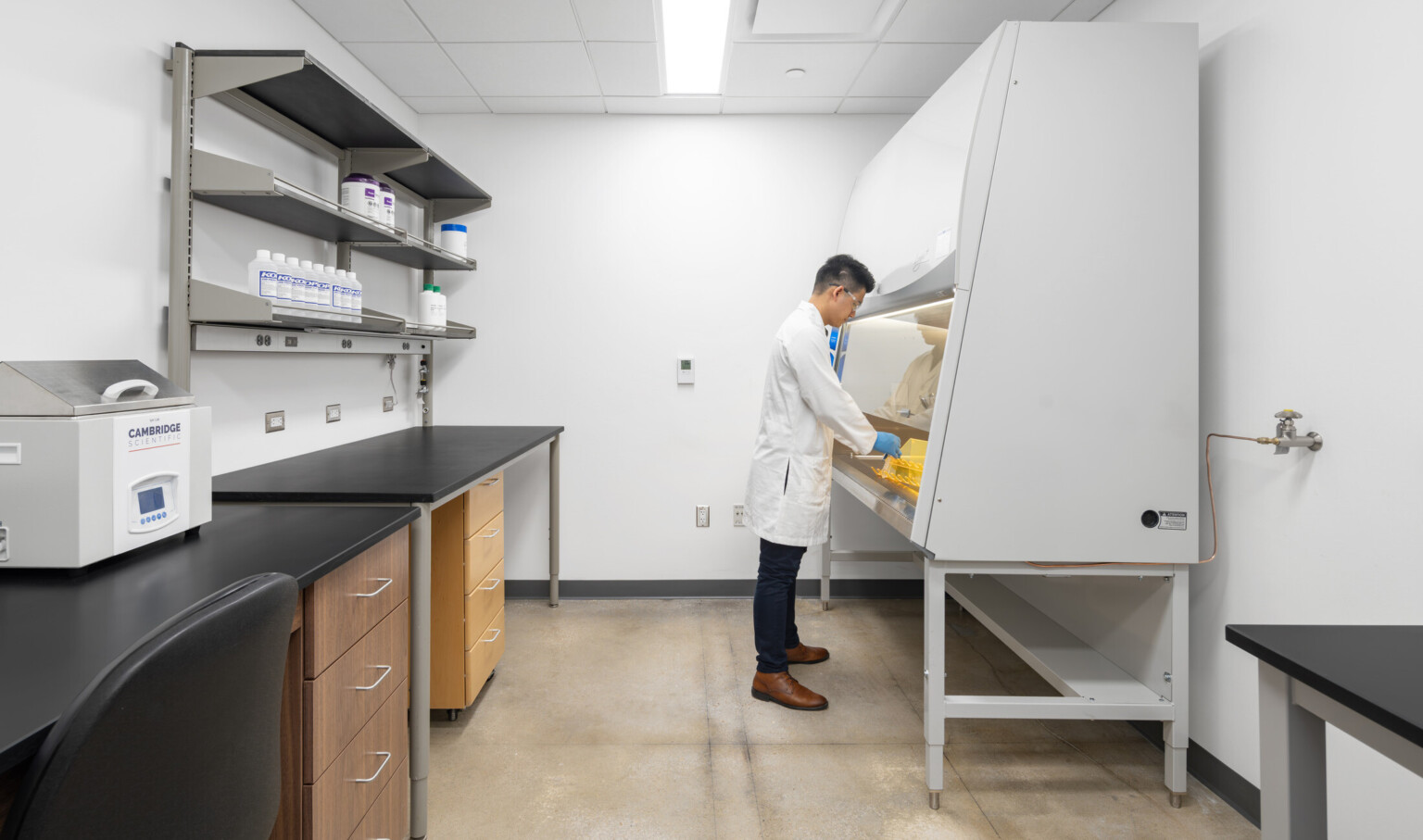 Person in a lab coat working at a fume hood. The lab includes shelves stocked with bottles and containers, a countertop with drawers, and equipment.