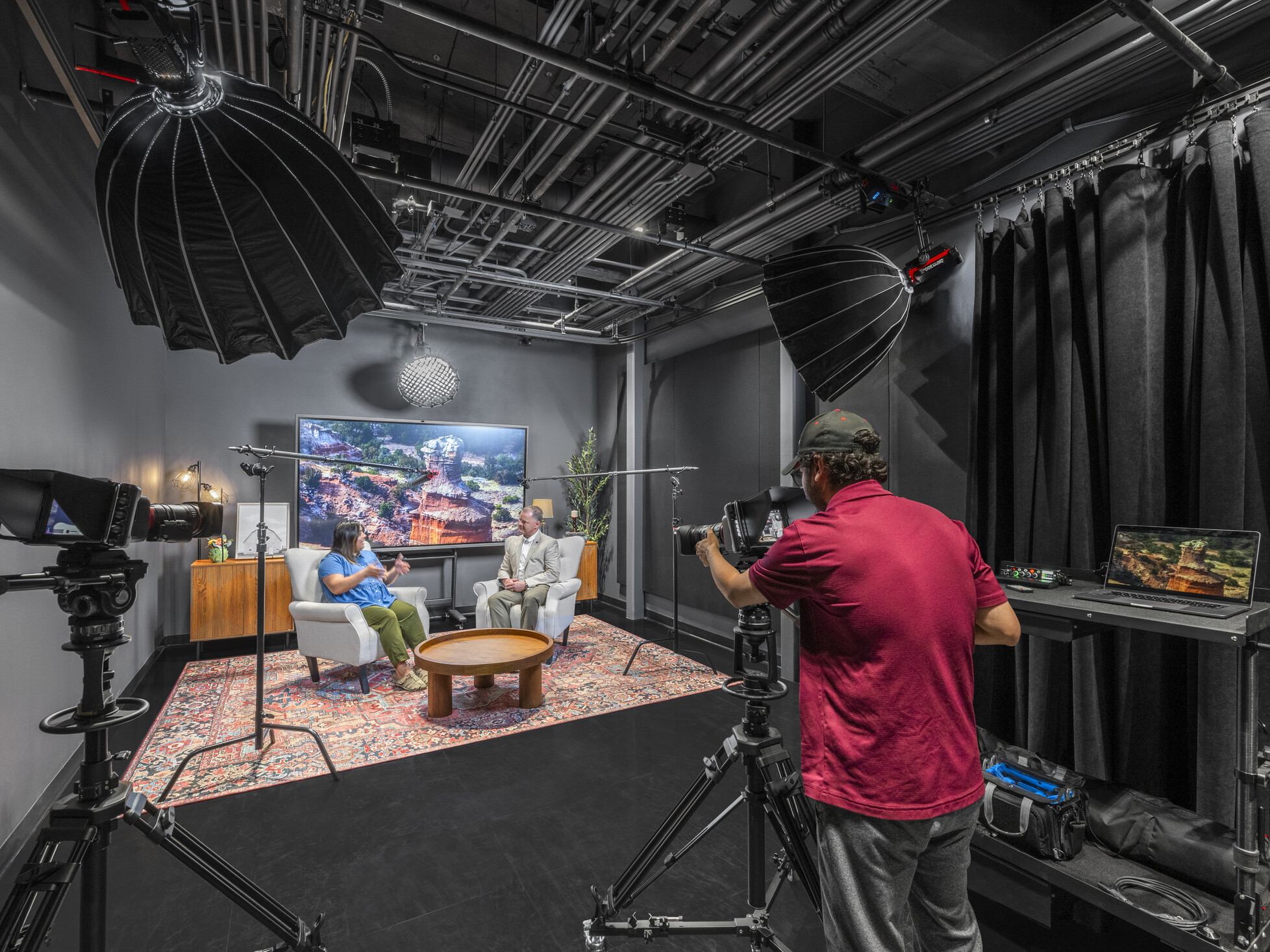 interior podcast filming room; modern tech surrounds a small set with two people talking to each other