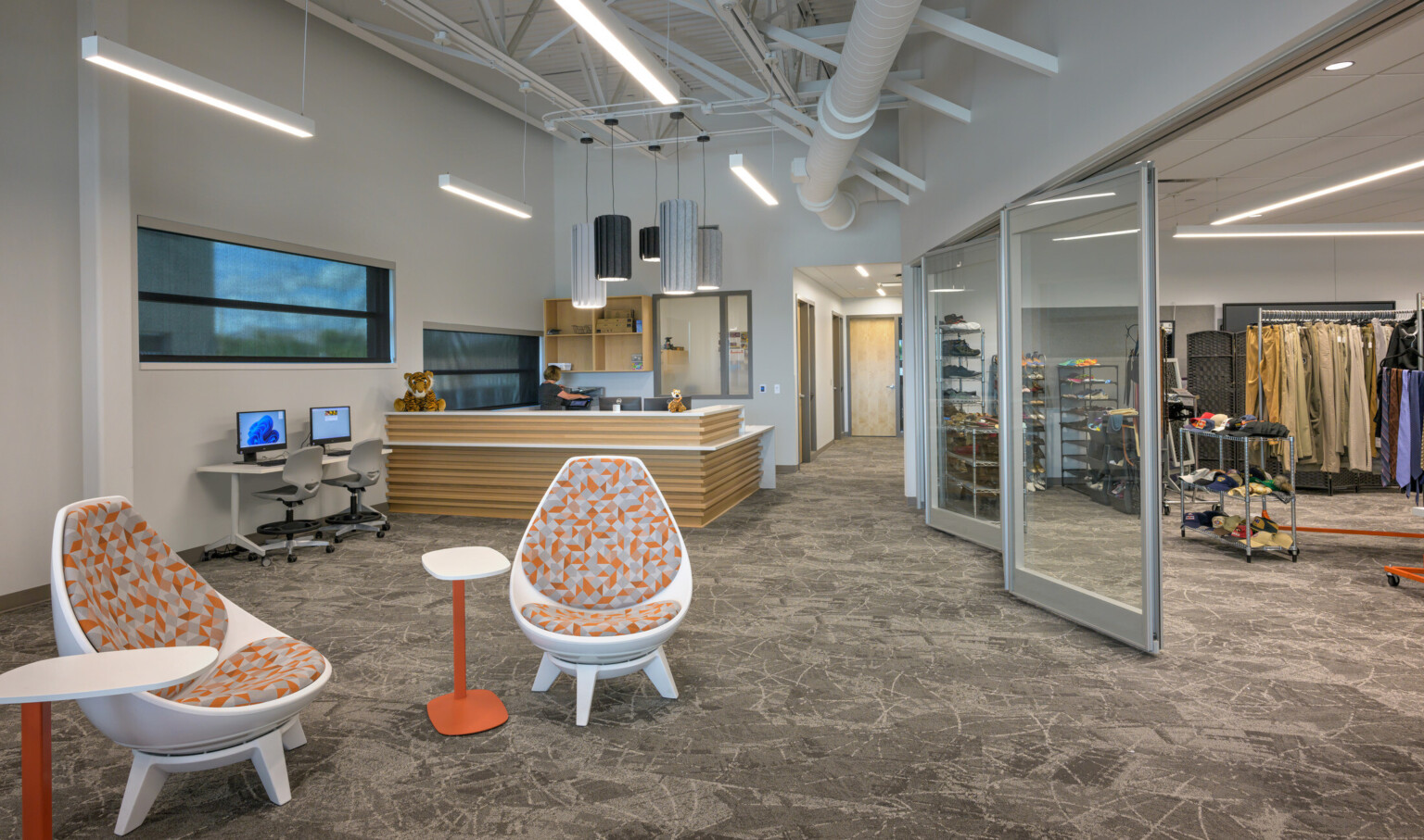 Multi-use school space with ergonomic chairs and tables in the foreground; an information desk and woman in the background with stuffed tigers; utility space to the right with closing and shoes
