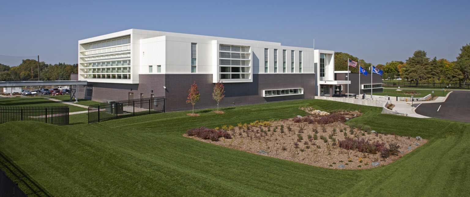 Multistory building with a dark brown bottom and white top surrounded by lush green grass with three flag flying along the concrete walkway