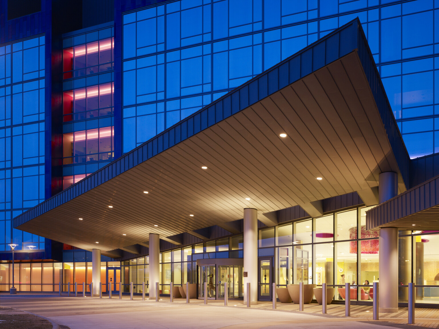 The front entrance of a modern medical building with large glass windows. A metal canopy supported by columns covers the entrance. Inside, warm yellow and orange lighting illuminates seating areas.