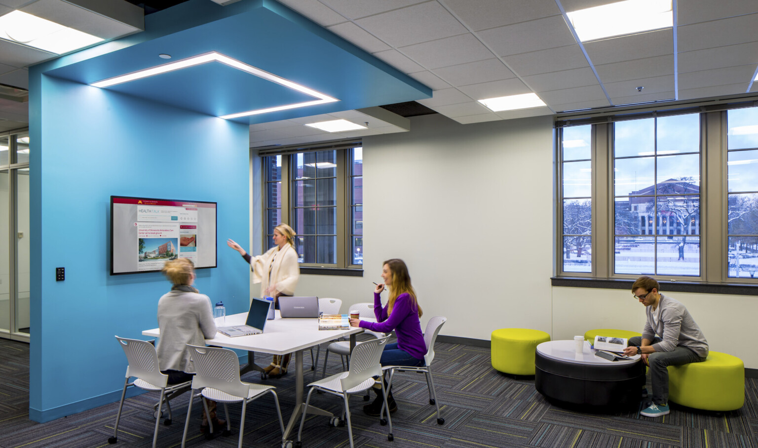 small group student work at a presentation screen under blue wrapped ceiling accent which goes to floor. Illuminated above small group student work at a presentation screen under blue wrapped ceiling accent which goes to floor. Illuminated above