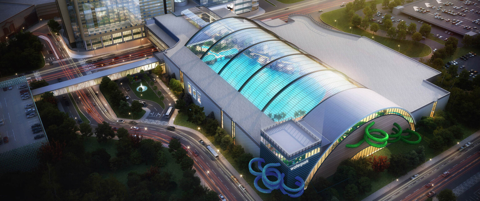Aerial view of indoor waterpark with arching skylights over roof. Blue and green water slides curl out sides of grey building